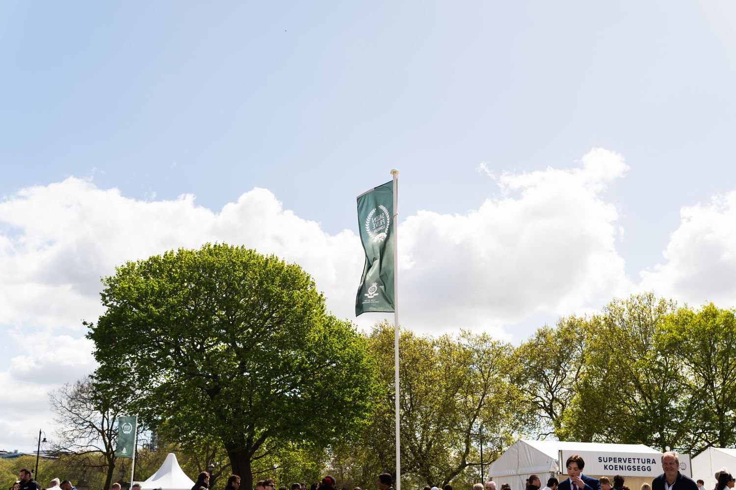 Green flag flying at an outdoor luxury automotive show with white tents and trees.