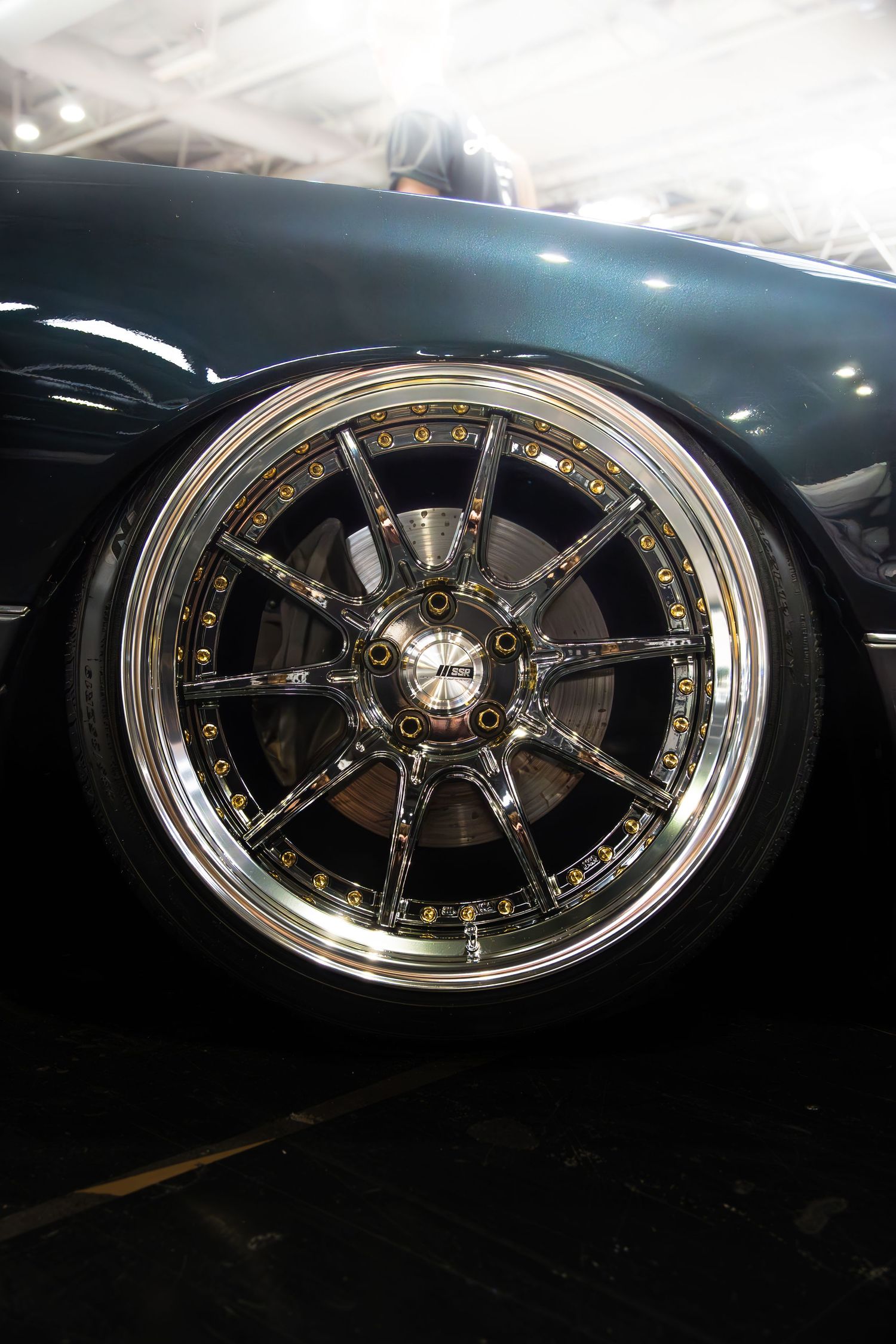 Chrome multi-spoke aftermarket alloy wheel with gold bolts on a black luxury car.