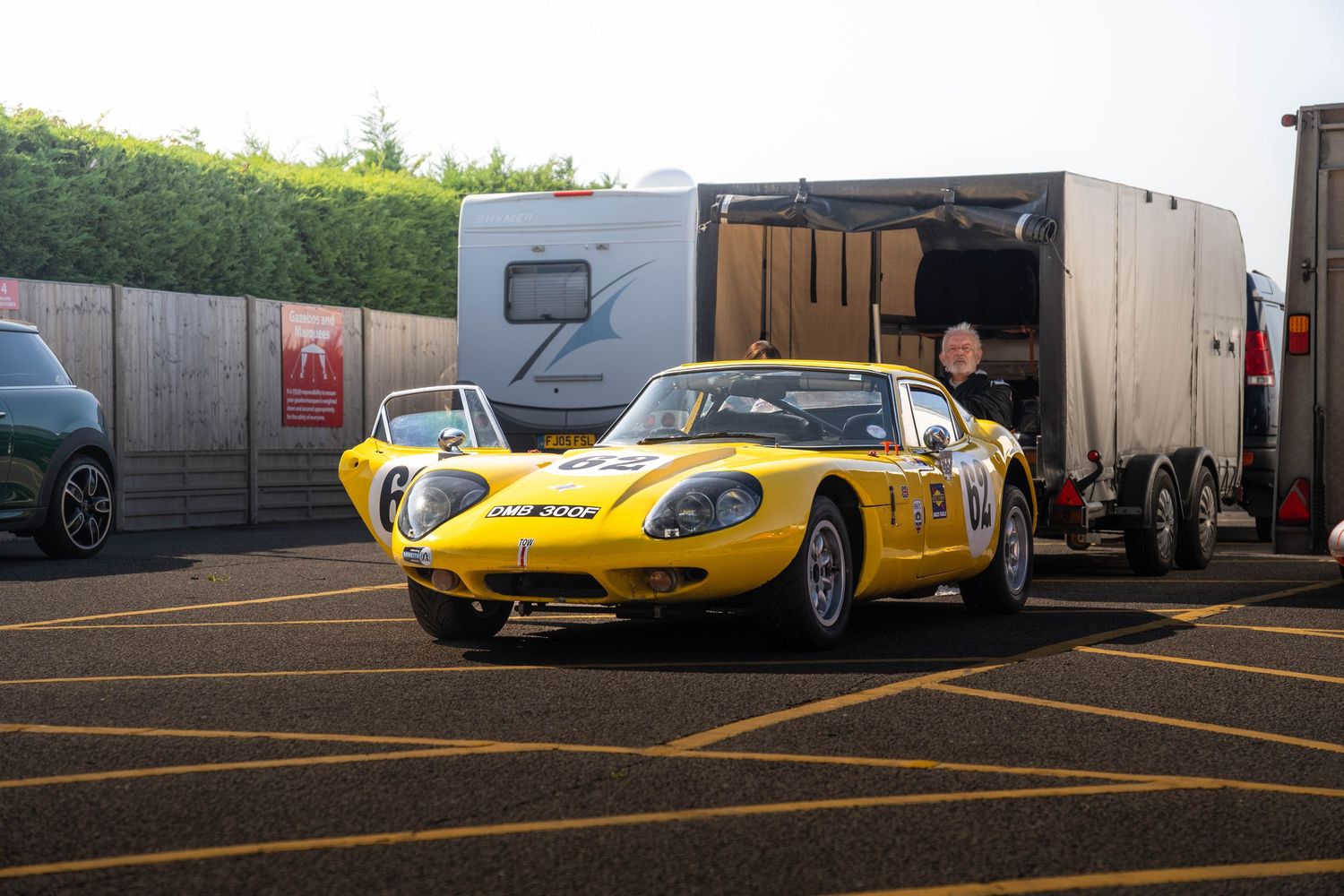Bright yellow vintage sports racing car parked in paddock area near transport trailer.