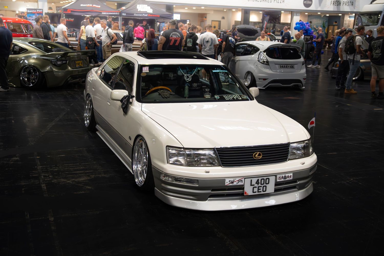 White lowered Lexus LS400 with custom body kit and chrome wheels at an indoor car show.
