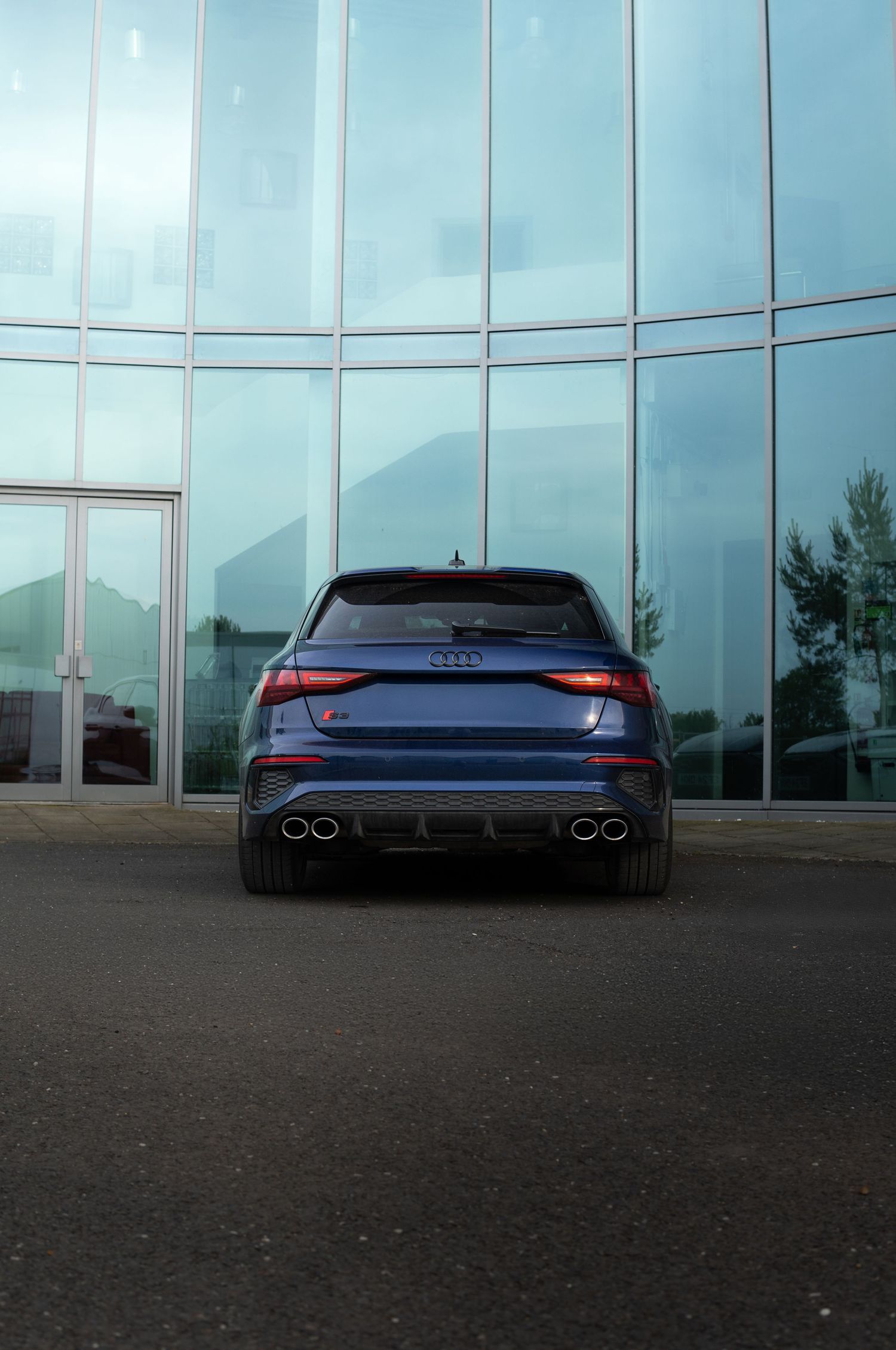Rear view of a navy blue Audi S3 with quad exhaust tips parked before a glass wall.