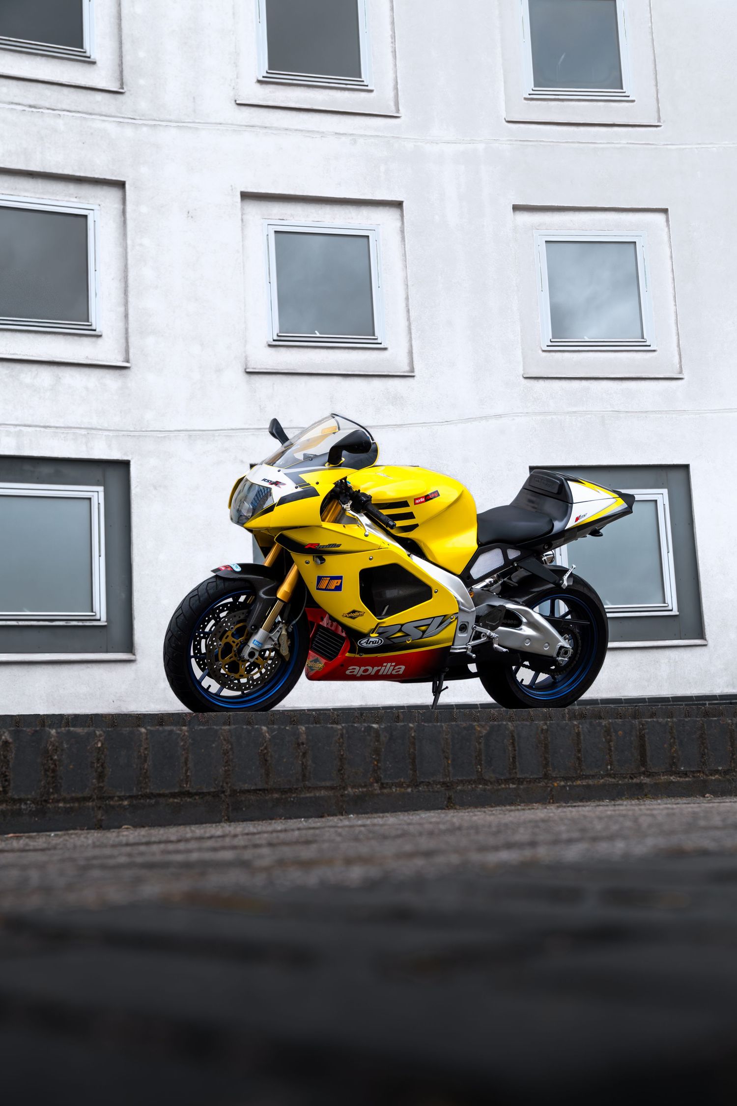 Yellow Aprilia RSV-R motorcycle parked on a raised curb against a grey paneled building wall.