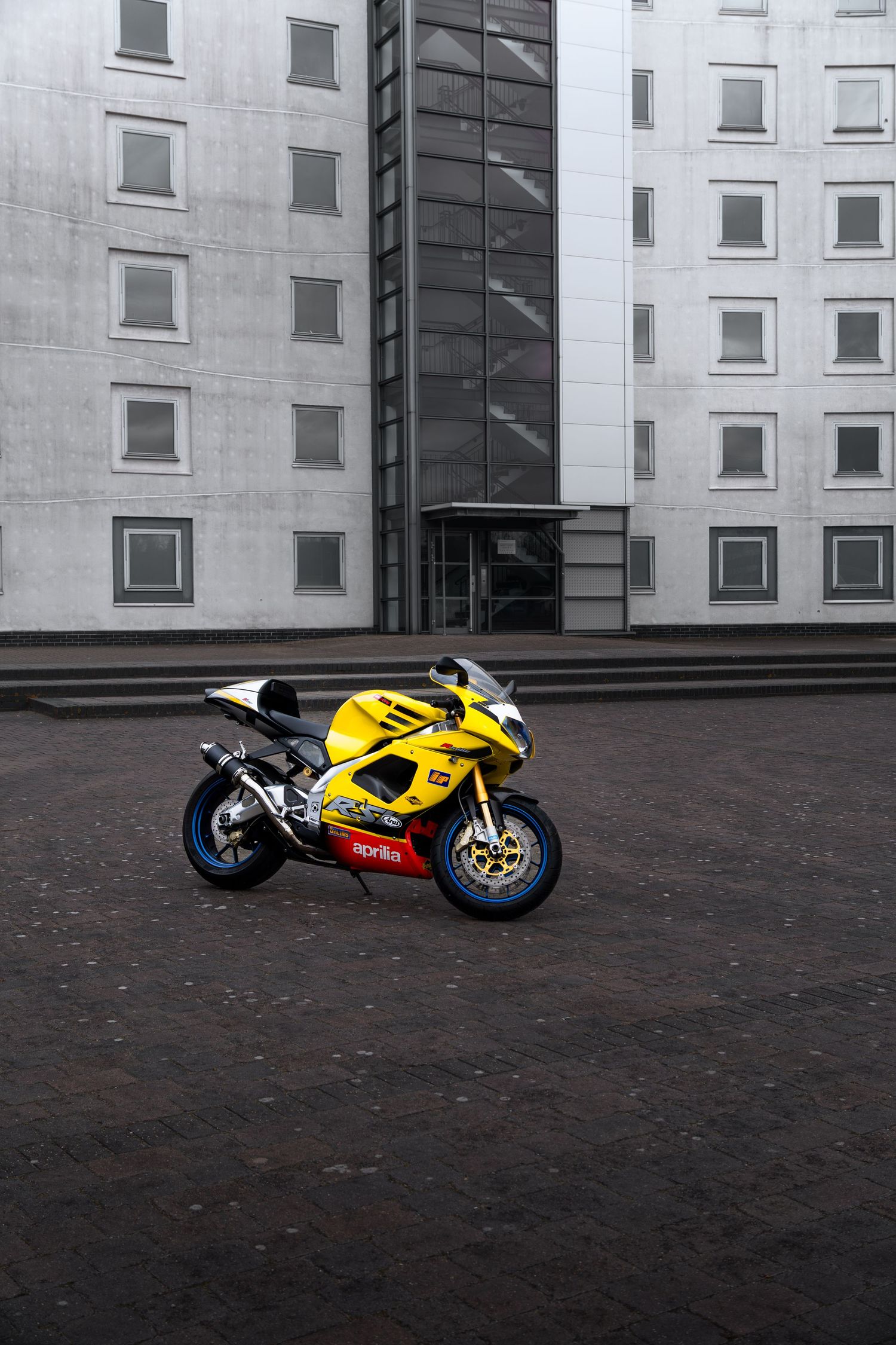 Yellow Aprilia RSV superbike on cobblestones with a tall modern white and glass building in background.