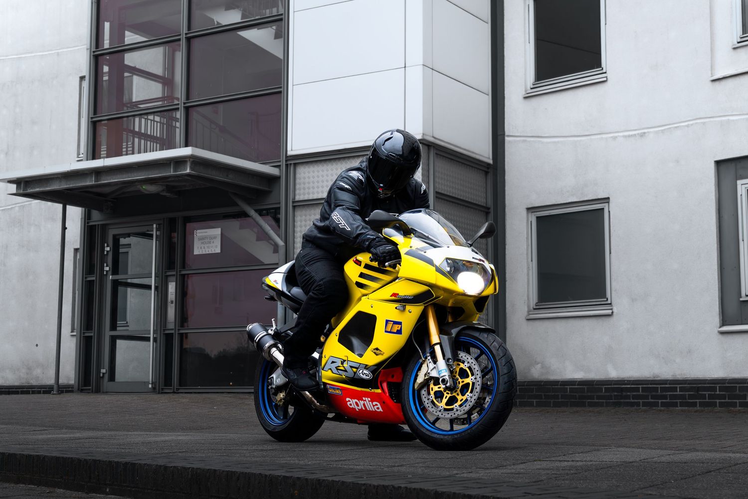 Yellow Aprilia RSV motorcycle with rider in black leathers outside modern office building.