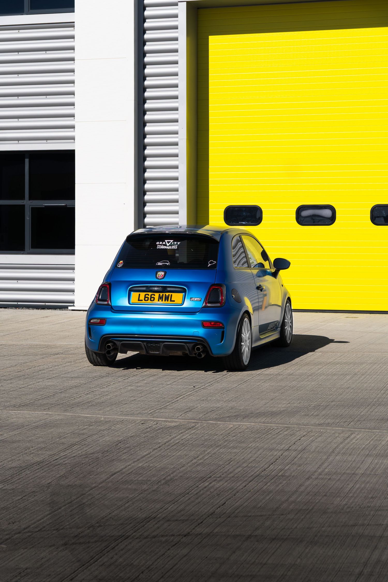 Three-quarter rear view of a blue Abarth 595 with diffuser beside yellow industrial garage doors.