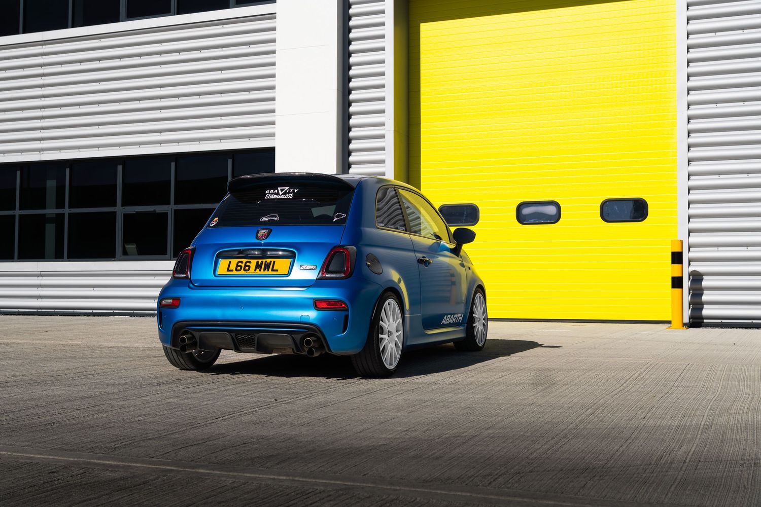 Rear view of a blue Abarth 595 parked in front of bright yellow industrial garage doors.