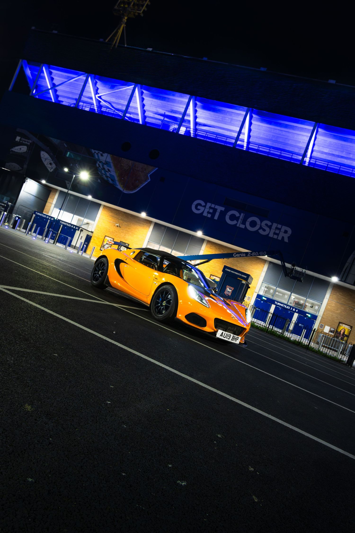 Vibrant orange Lotus Elise photographed at night outside illuminated blue stadium entrance with GET CLOSER sign.
