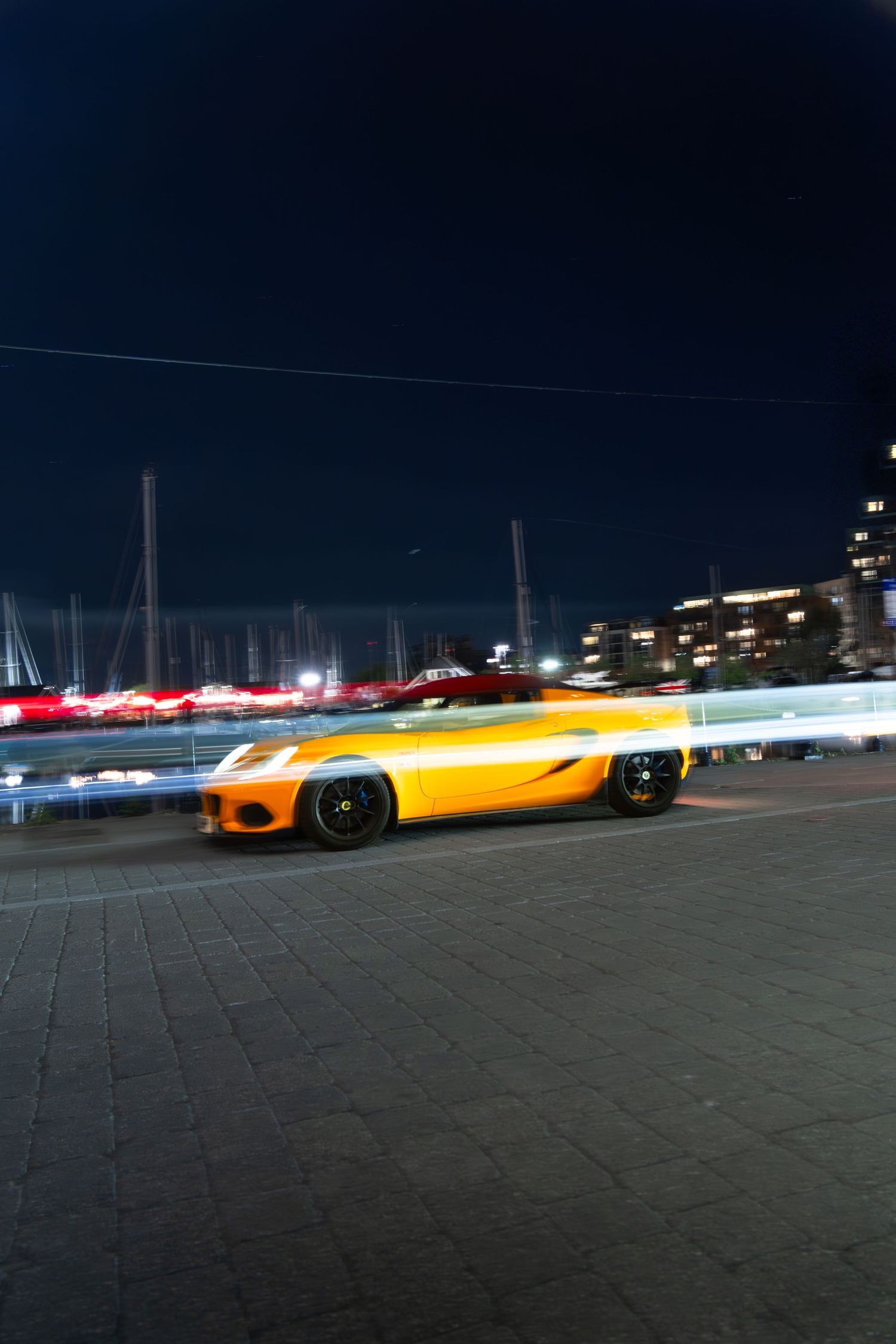 Yellow supercar captured with motion blur driving through illuminated waterfront area at night in city.