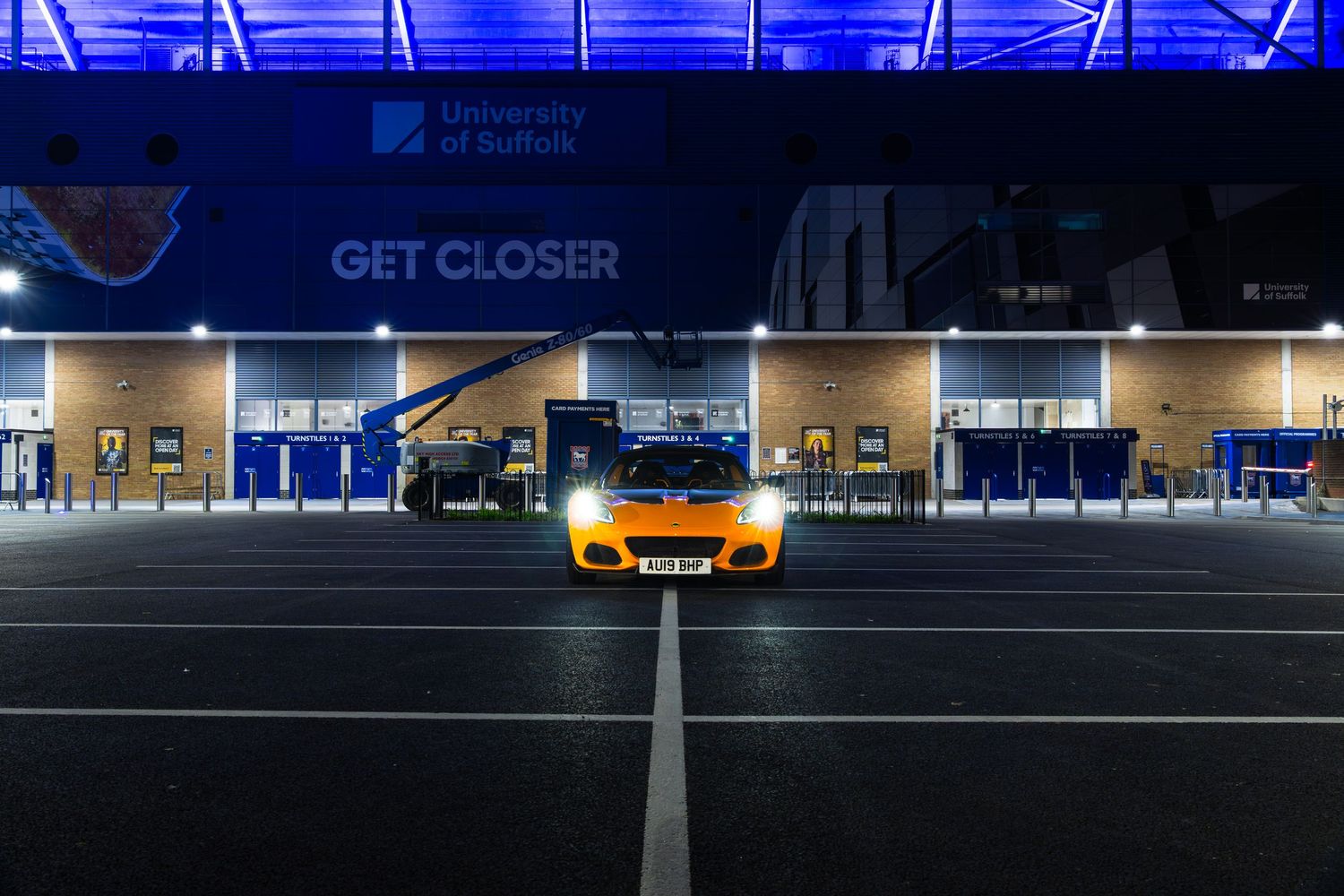 Orange Lotus Elise parked outside University of Suffolk GET CLOSER stadium at night with blue lighting.