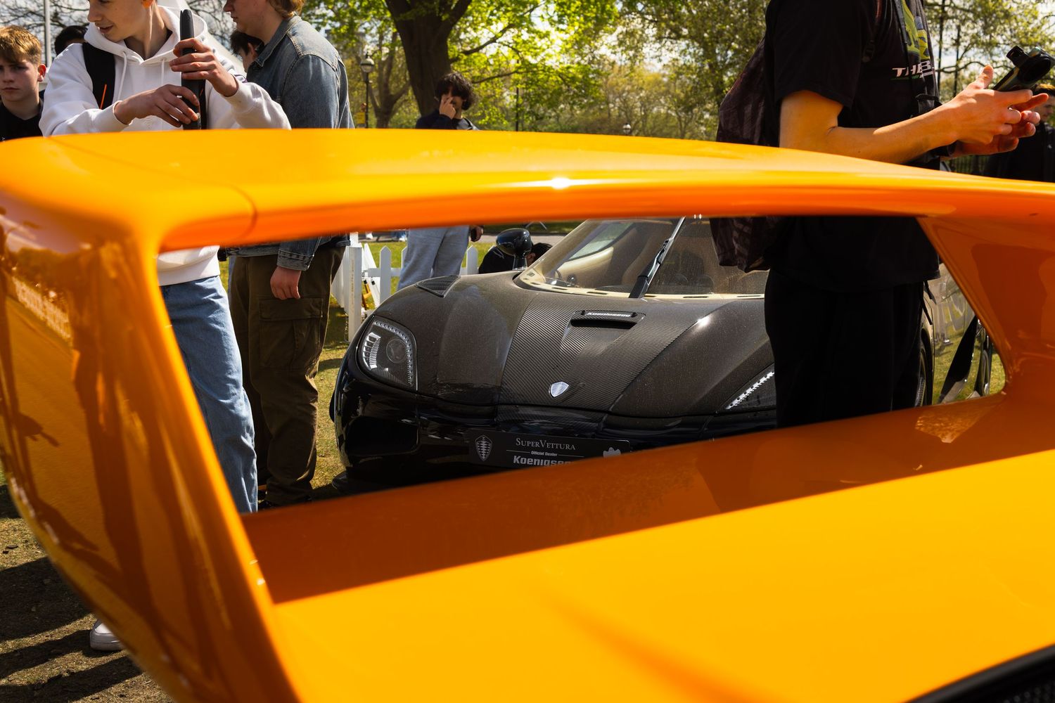 Orange supercar exterior at an outdoor exotic car show with spectators viewing a black hypercar through the window.