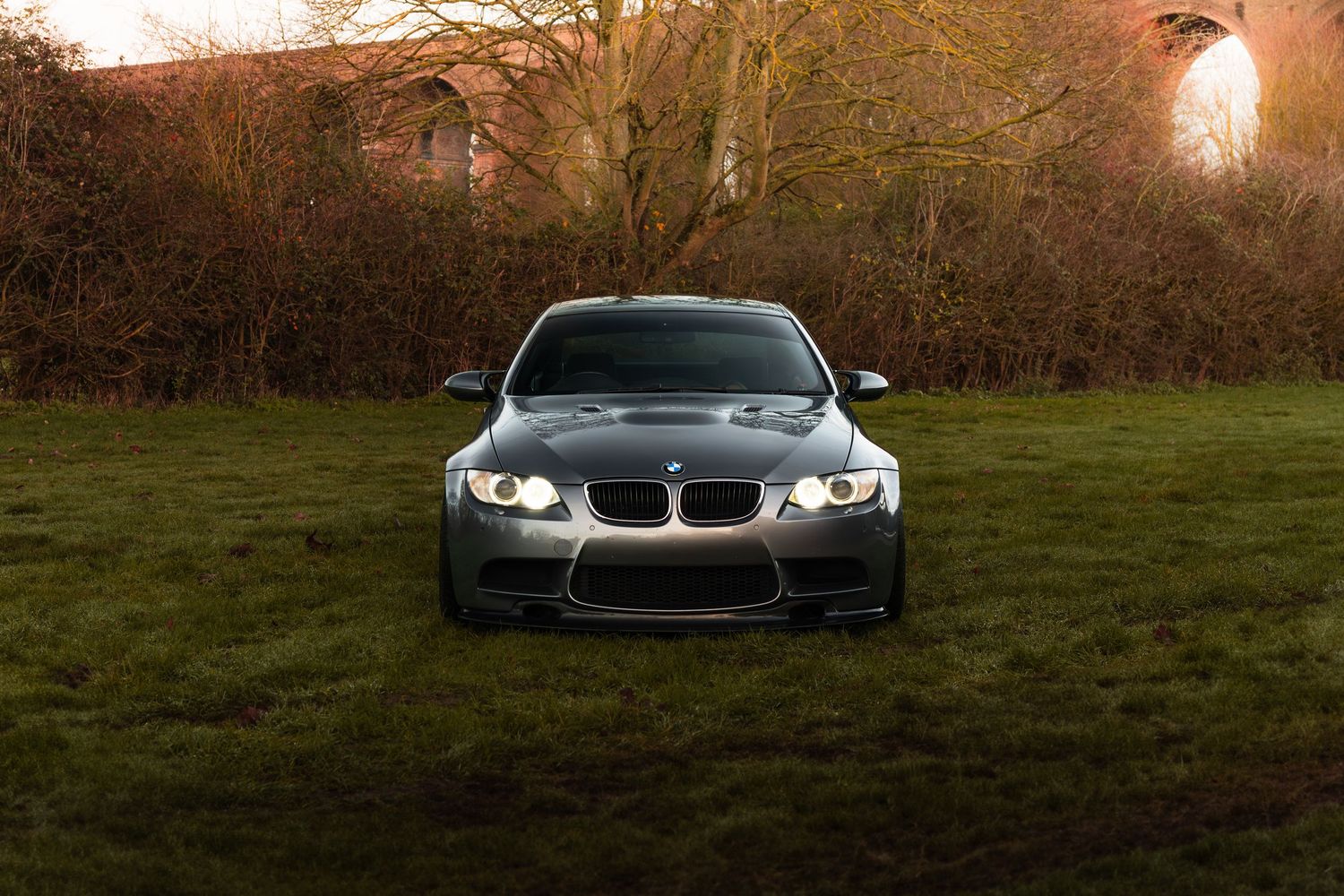 Front-facing silver BMW M3 with angel eye headlights parked on grass near brick arches.