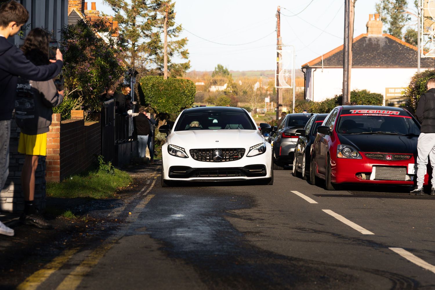 White Mercedes-AMG C63 leading a line of cars through a village street car meet.