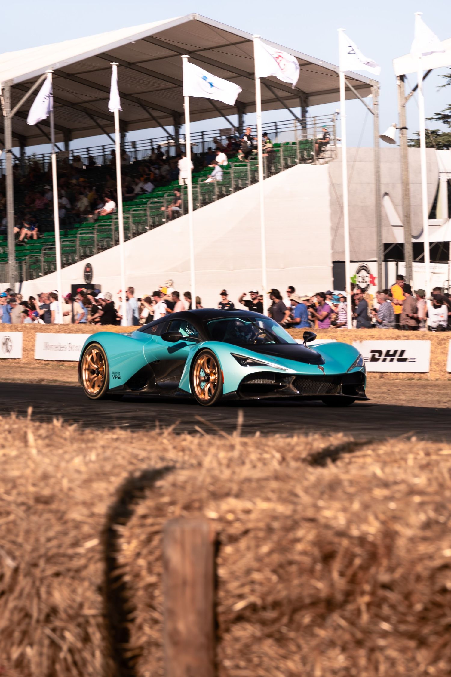 Teal supercar with gold wheels races at Goodwood Festival of Speed, crowds watching from grandstands behind hay bales.
