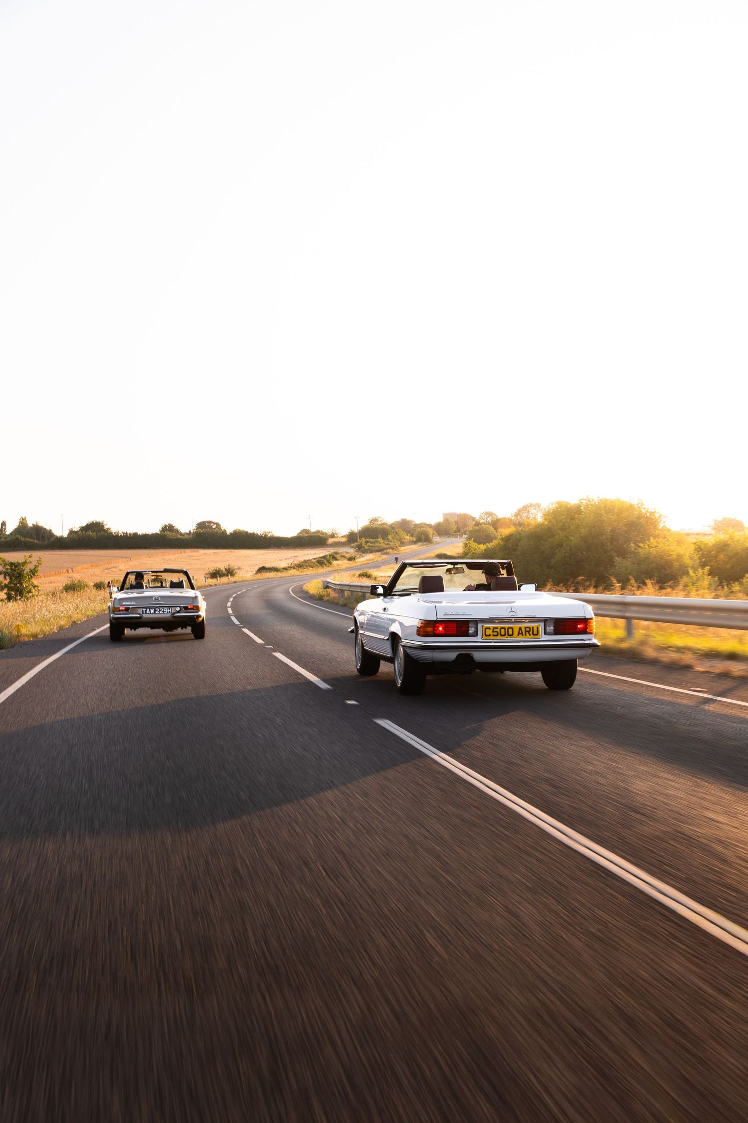 Two classic convertible cars driving on a scenic road at golden hour sunset, surrounded by fields and trees.