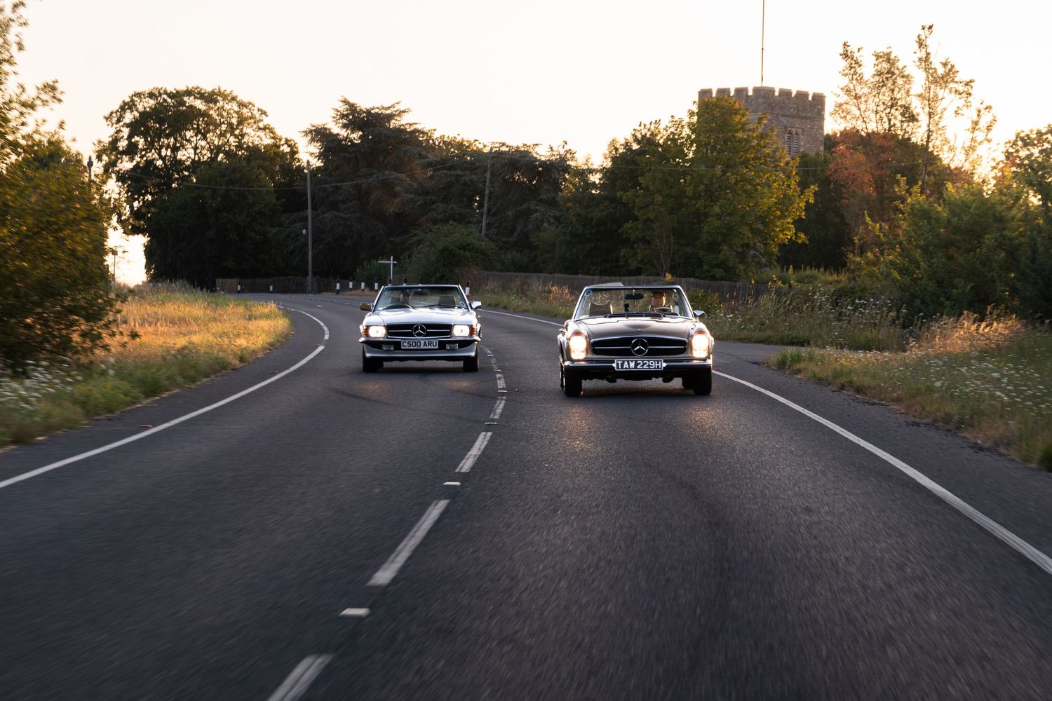 Two classic white cars driving on a rural road at dusk with trees and a stone tower in the background.