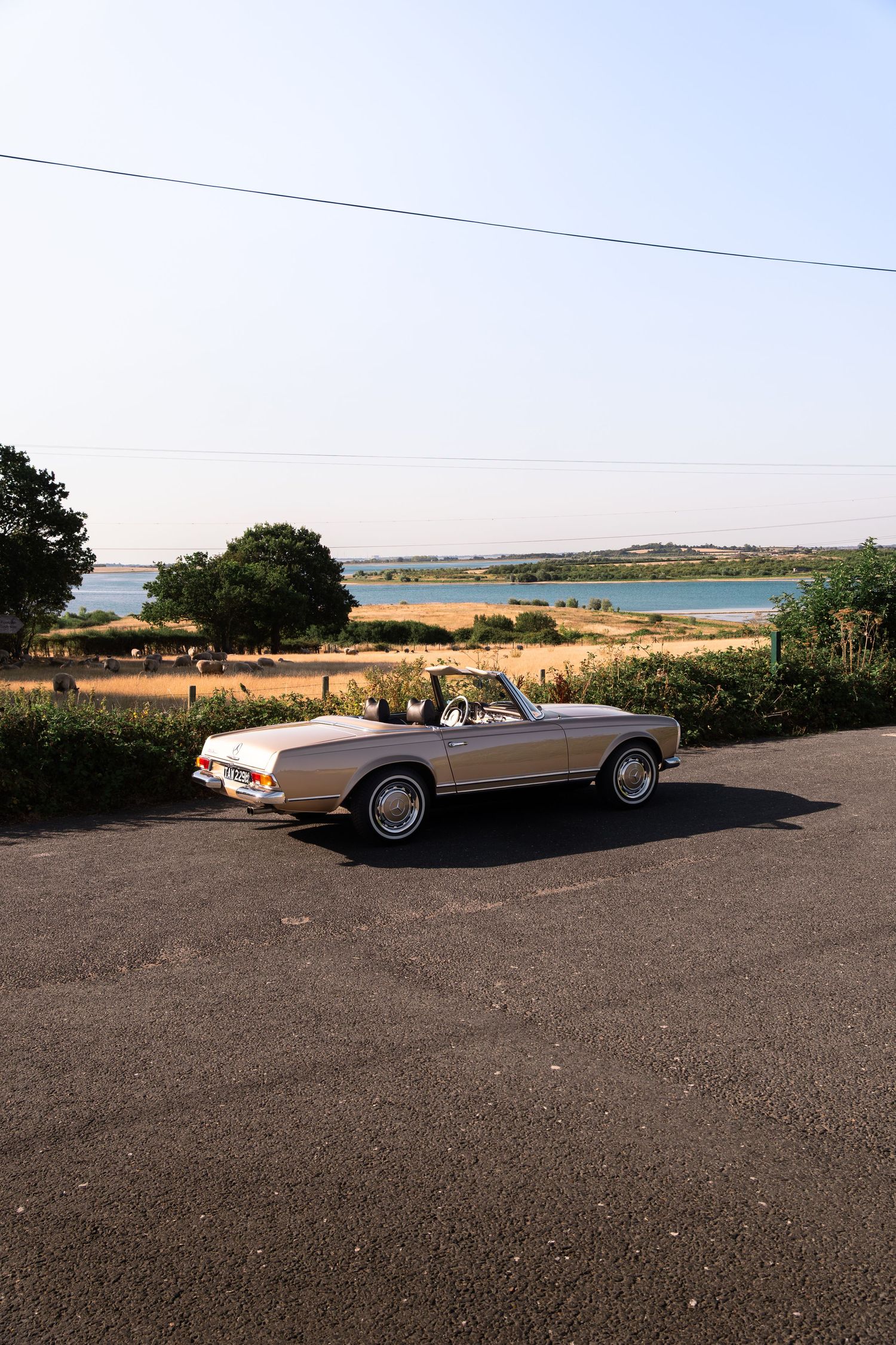 Vintage silver convertible car parked near a scenic coastal landscape with golden fields and blue water on a sunny day.
