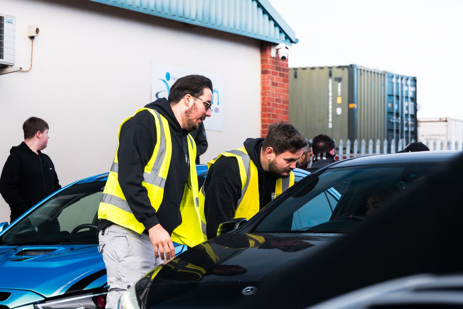 Two men in yellow safety vests inspecting a blue sports car at an outdoor car event.