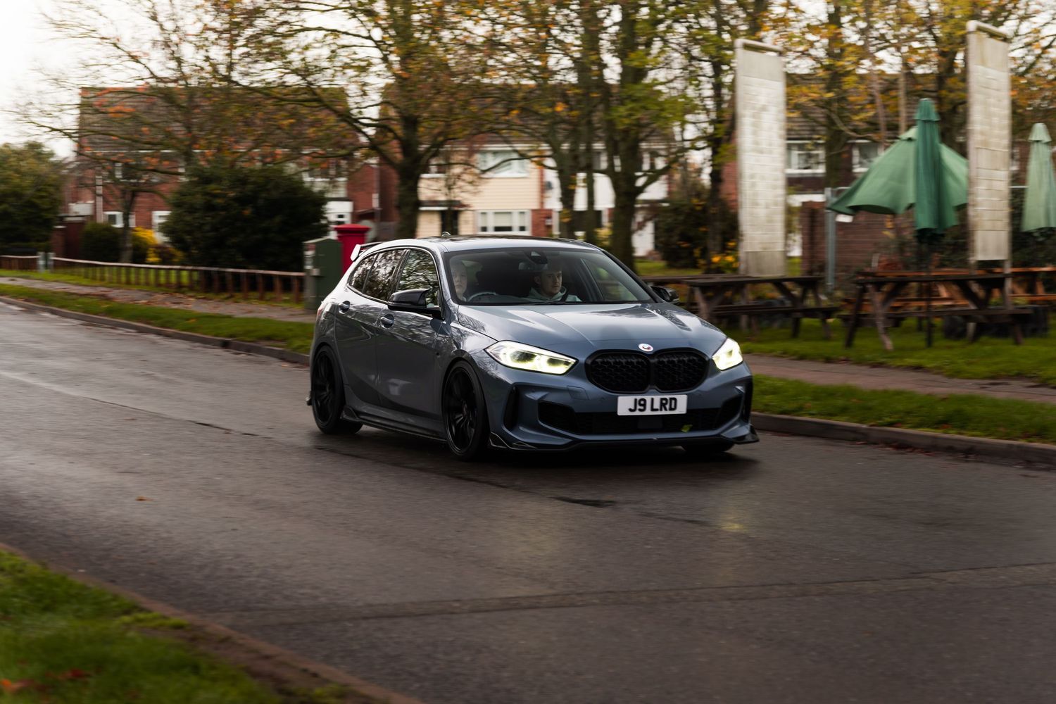 Dark grey BMW 1 Series hatchback driving on wet suburban road.