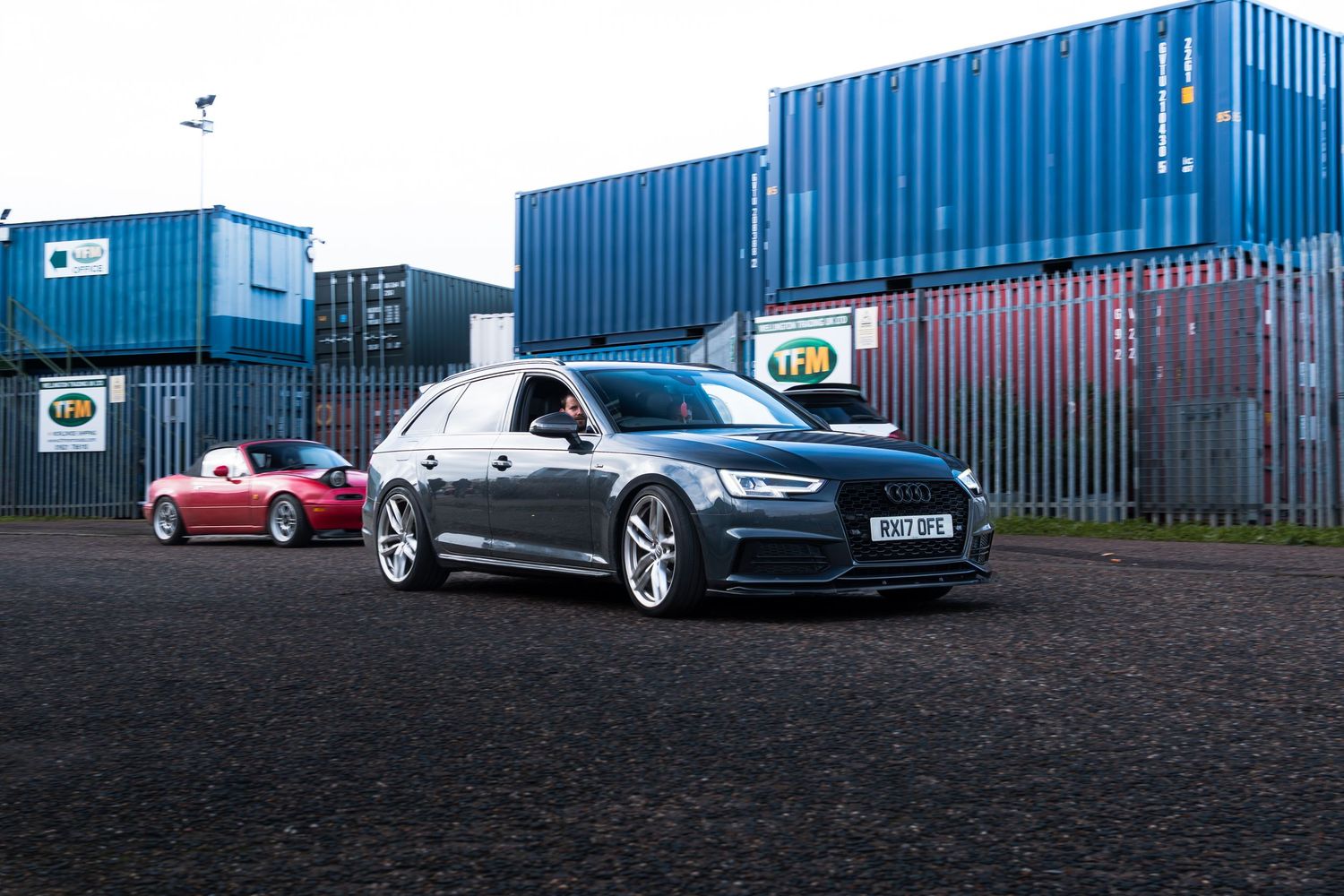 Grey Audi A6 Avant estate parked in front of blue shipping containers outdoors.