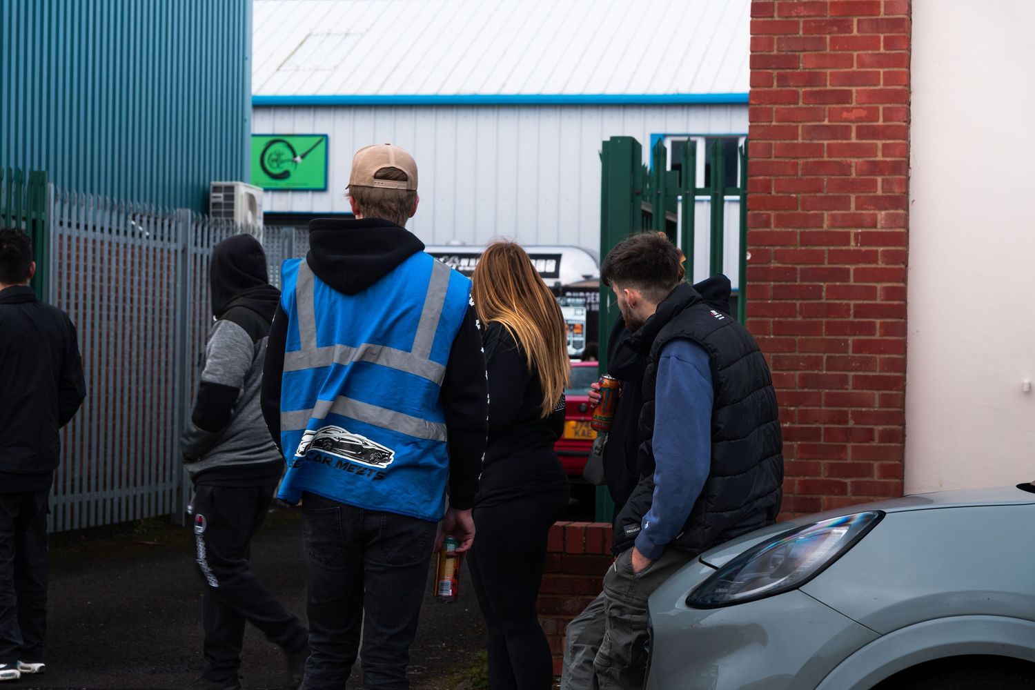 A group of people including a hi-vis worker gather outside a blue industrial building at a car event.