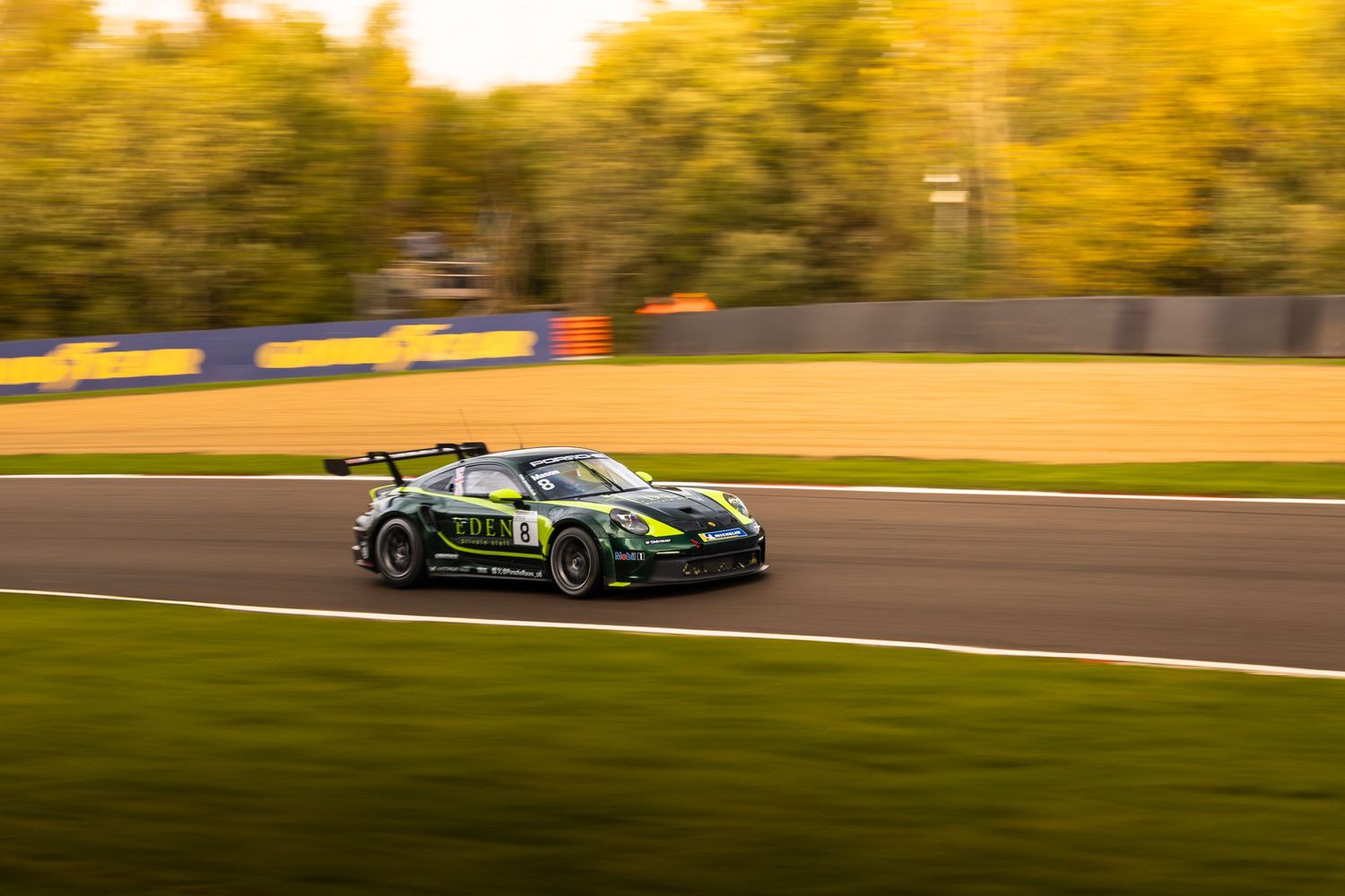 Dark green Porsche GT3 race car panning shot on racetrack with golden autumn trees background.