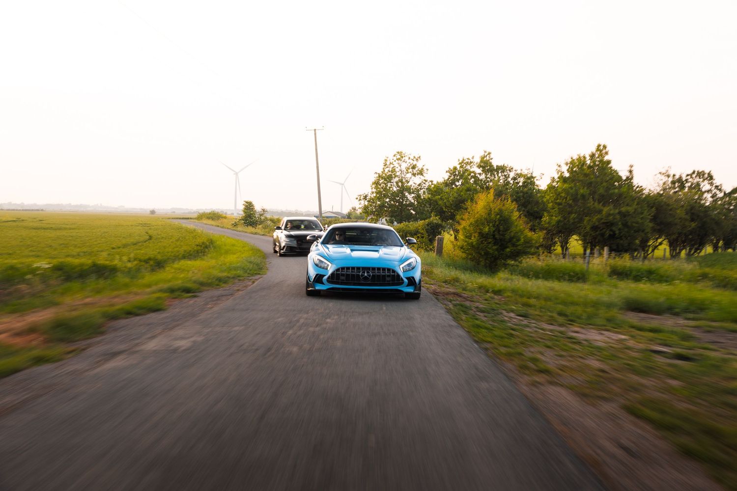 Two sports cars including a blue Mercedes-AMG GT cruising on a rural road at golden hour.