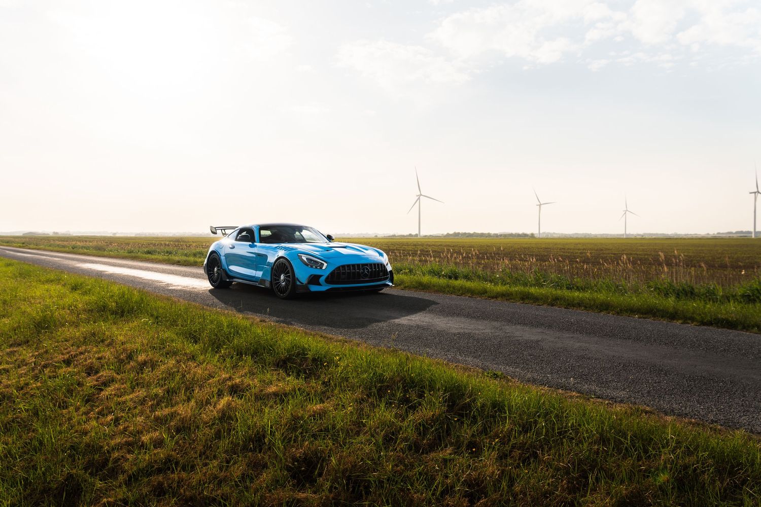 Blue Mercedes-AMG GT Black Series driving on straight rural road with wind turbines.
