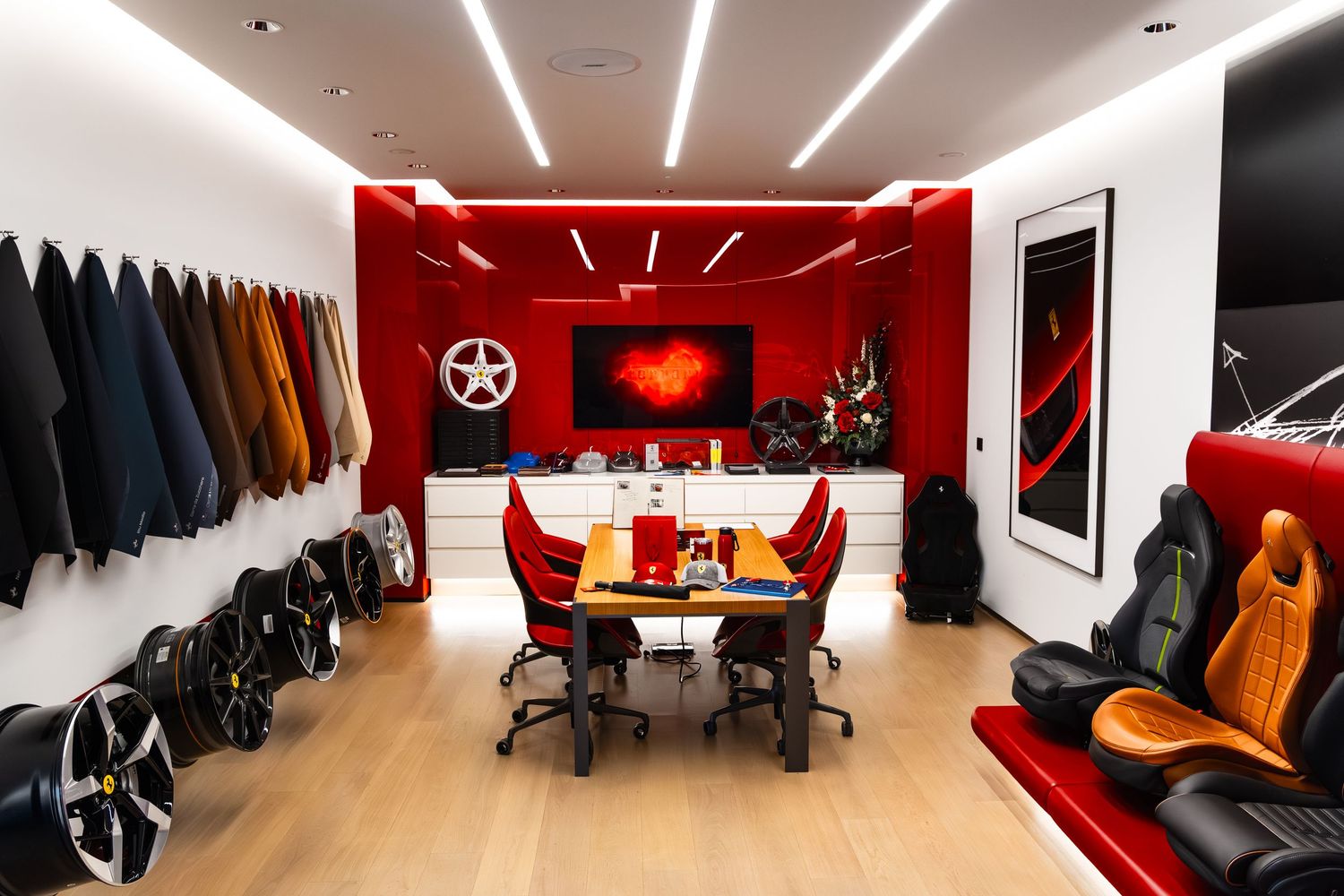 Ferrari showroom interior with red accent wall, wheel displays, leather seat samples, and a central meeting table on hardwood floors.