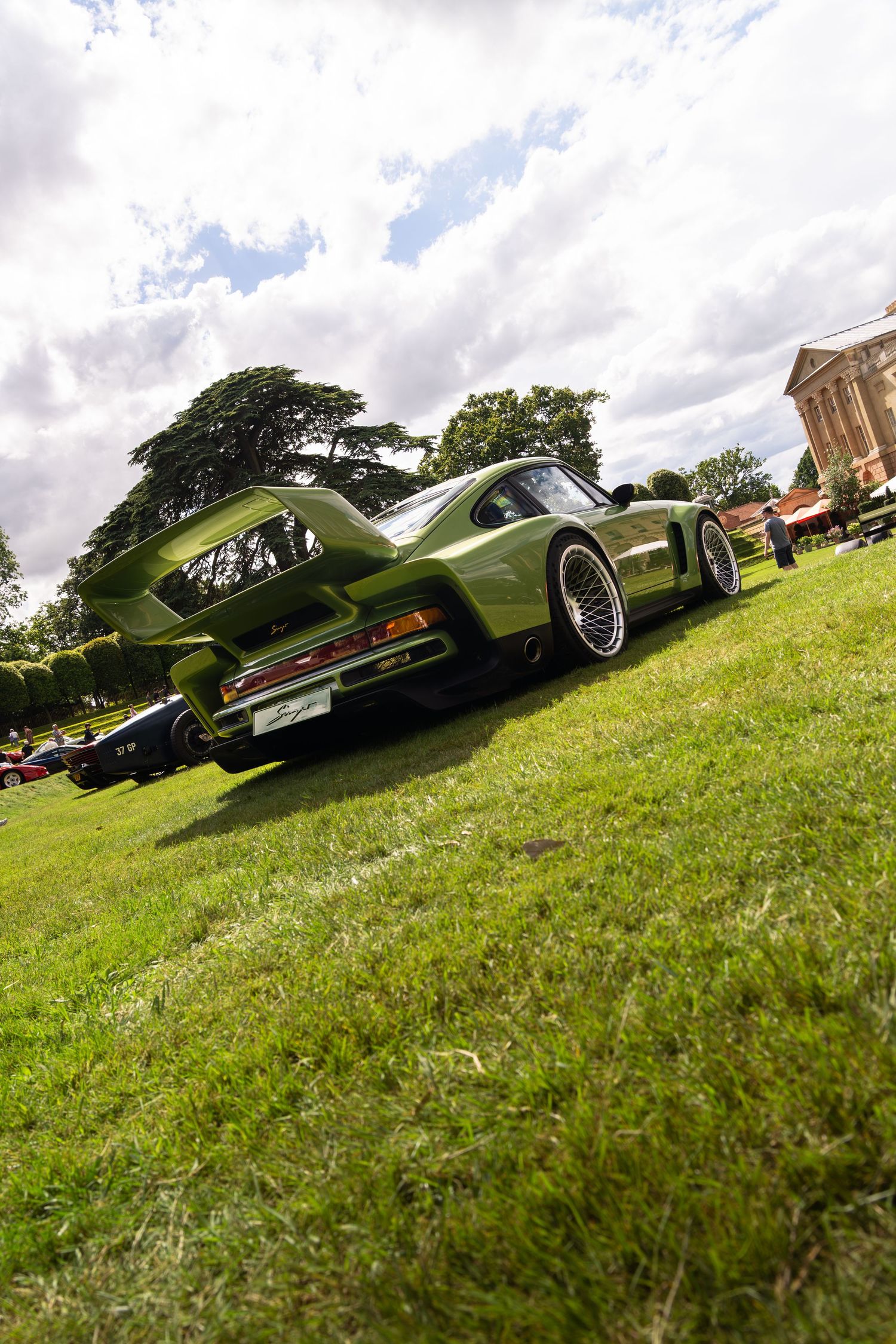 Olive green Porsche sports car displayed on grass at an outdoor classic car show under a cloudy sky.