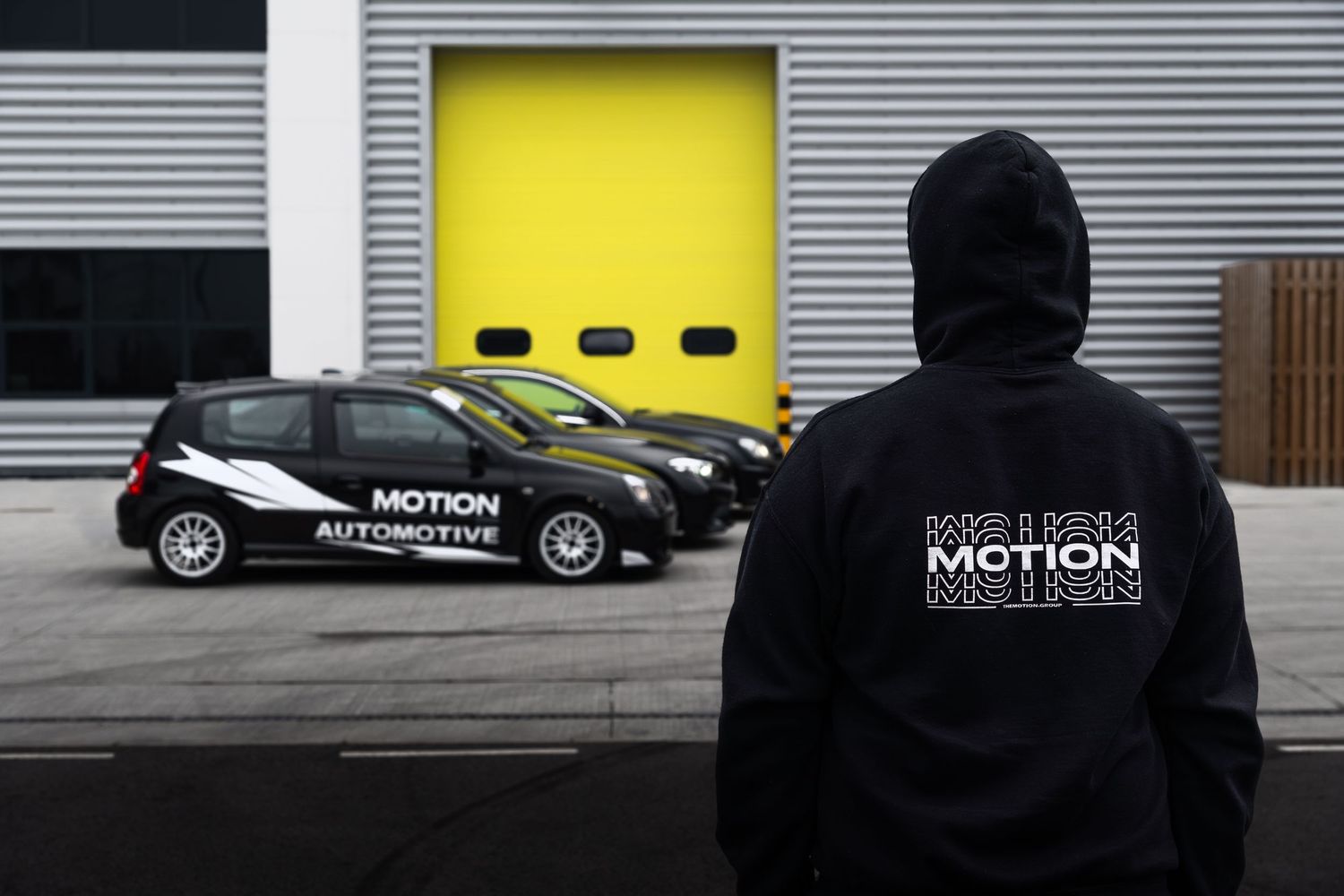 Person wearing a Motion Automotive hoodie standing before two branded black Renault vehicles outside a yellow garage door.