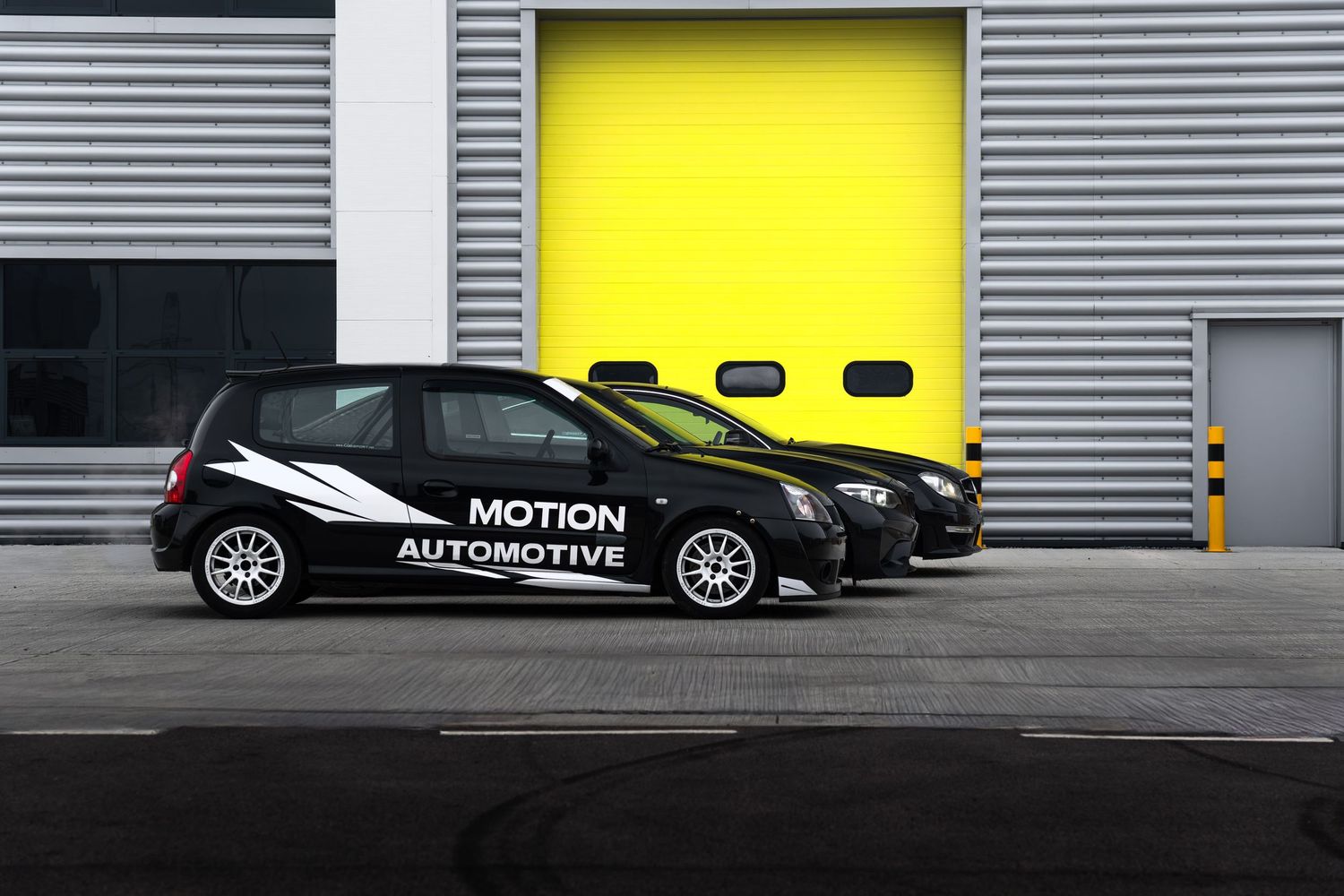Three black Motion Automotive branded cars parked in front of a bright yellow industrial roller door.