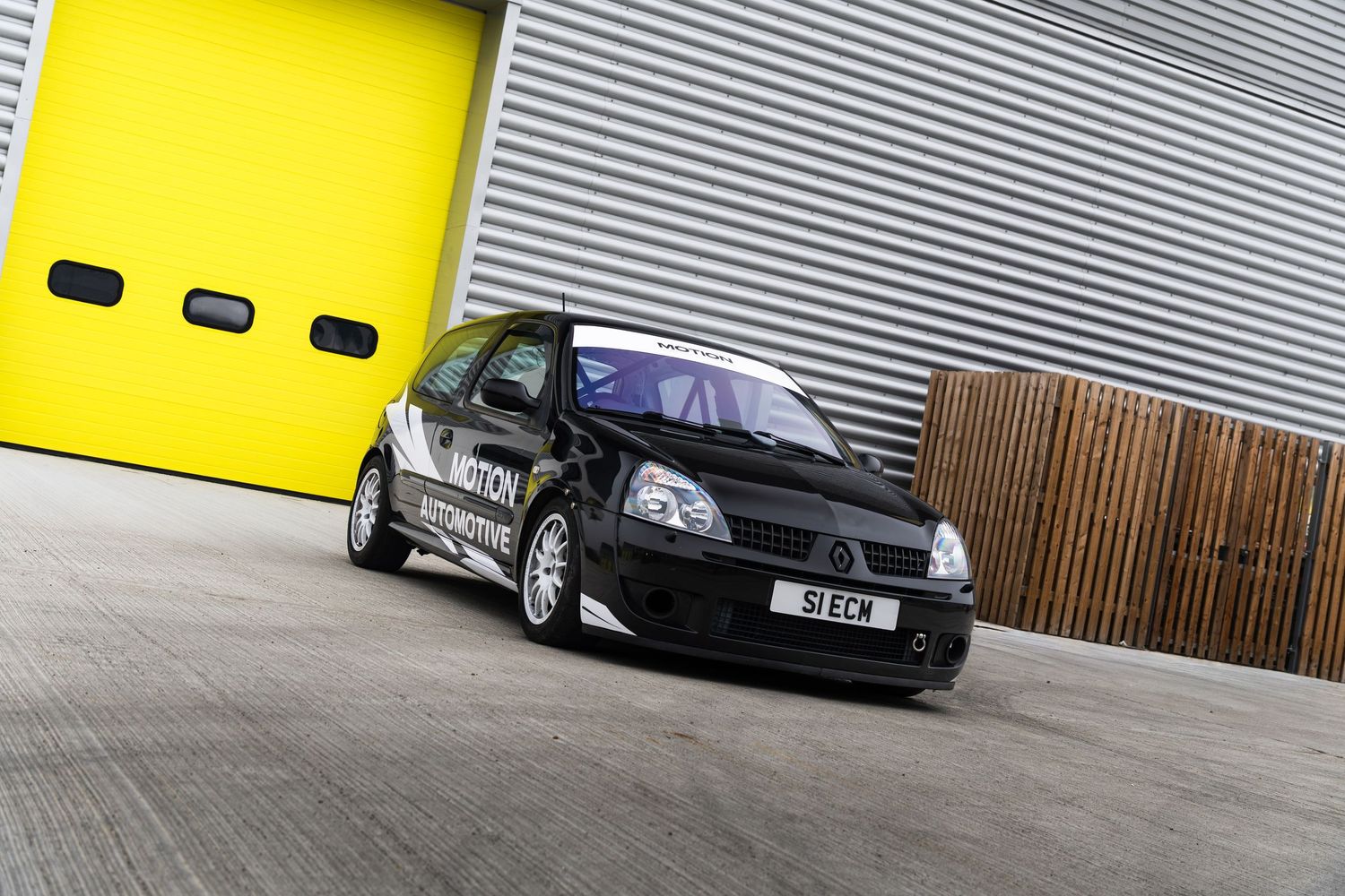 Black Renault Clio with Motion motorsport livery parked outside industrial garage with yellow doors.