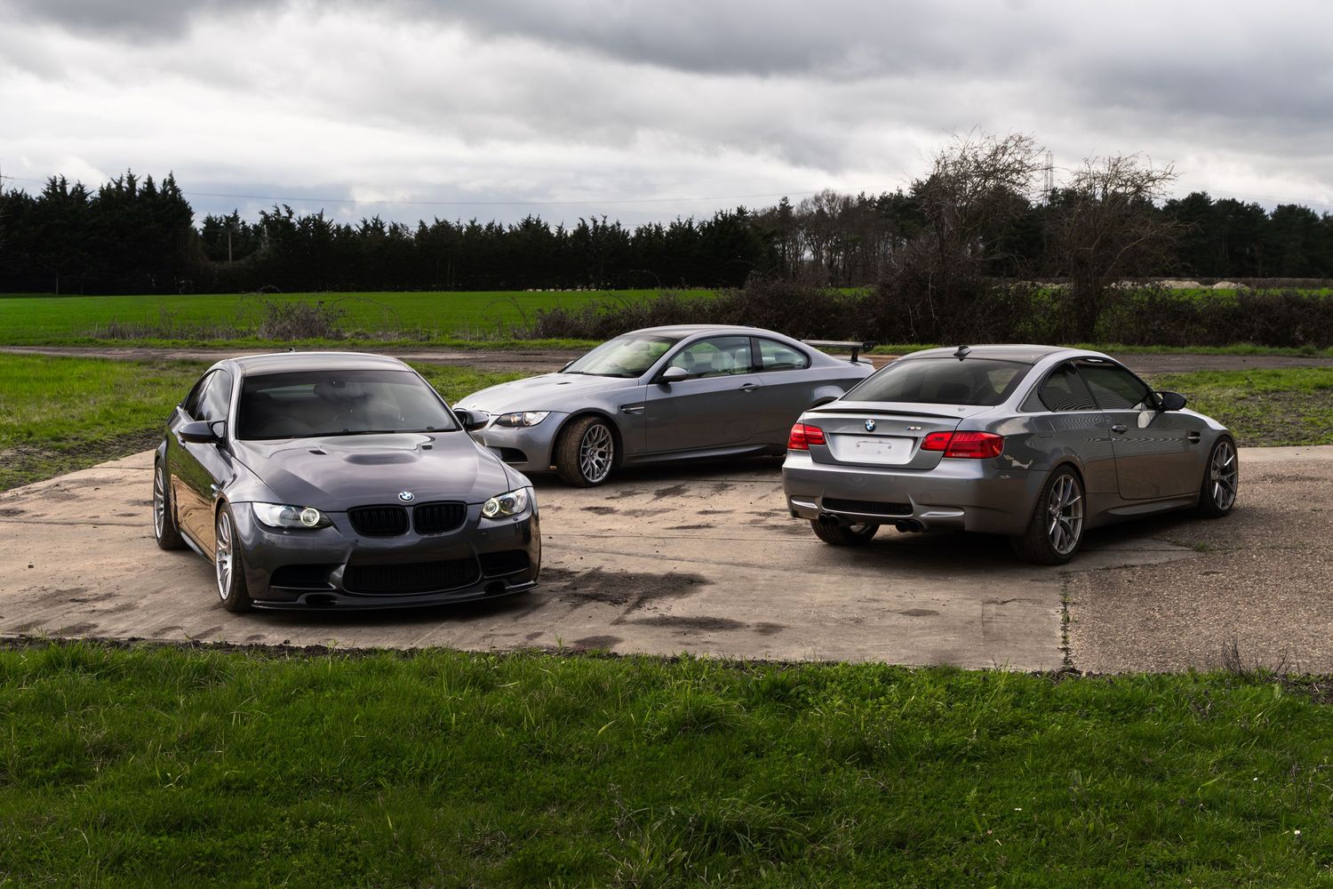 Three gray BMW M3 coupes parked on gravel surrounded by green fields under cloudy skies.