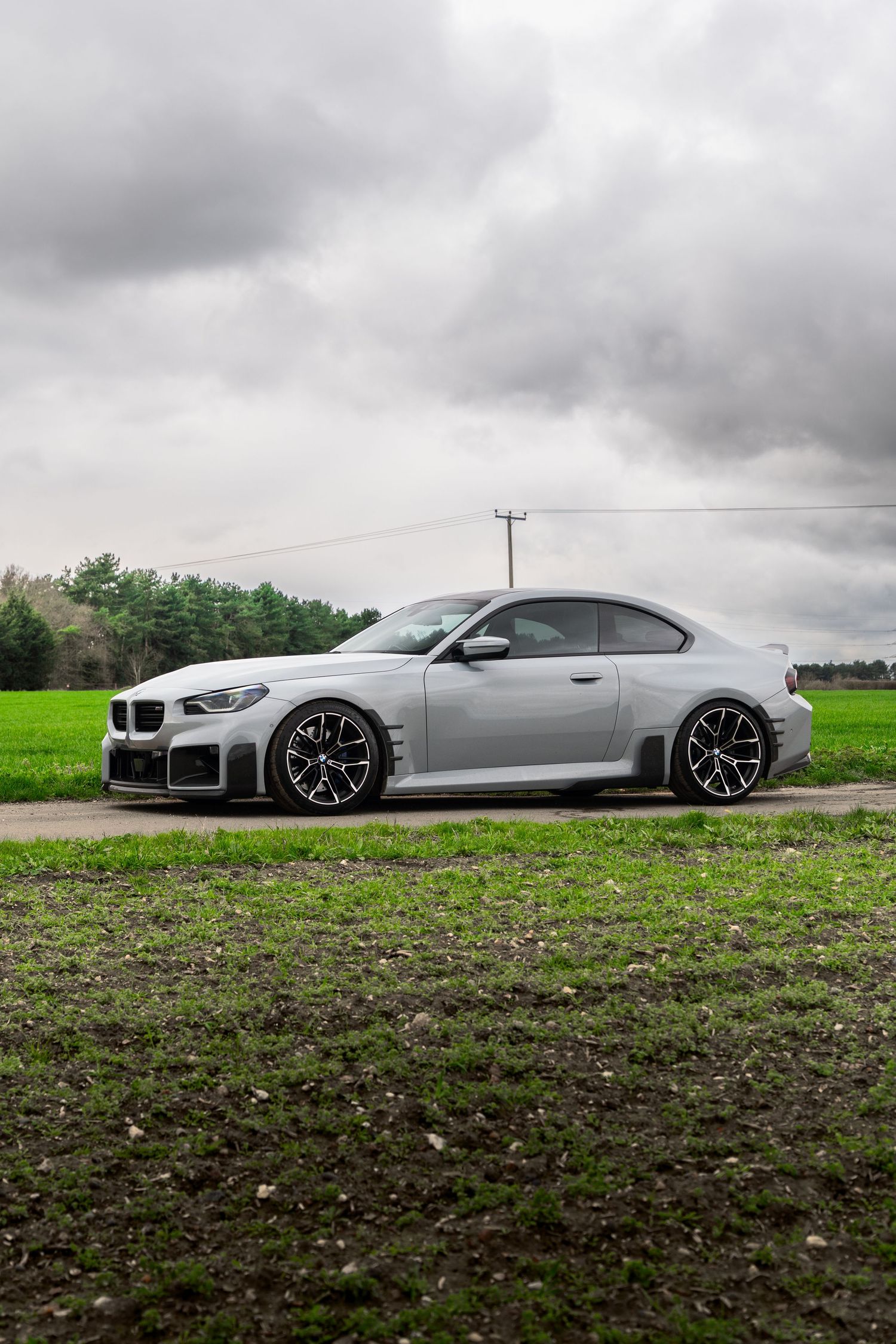Side view of a modified silver BMW M2 Competition coupe parked on a rural gravel road.
