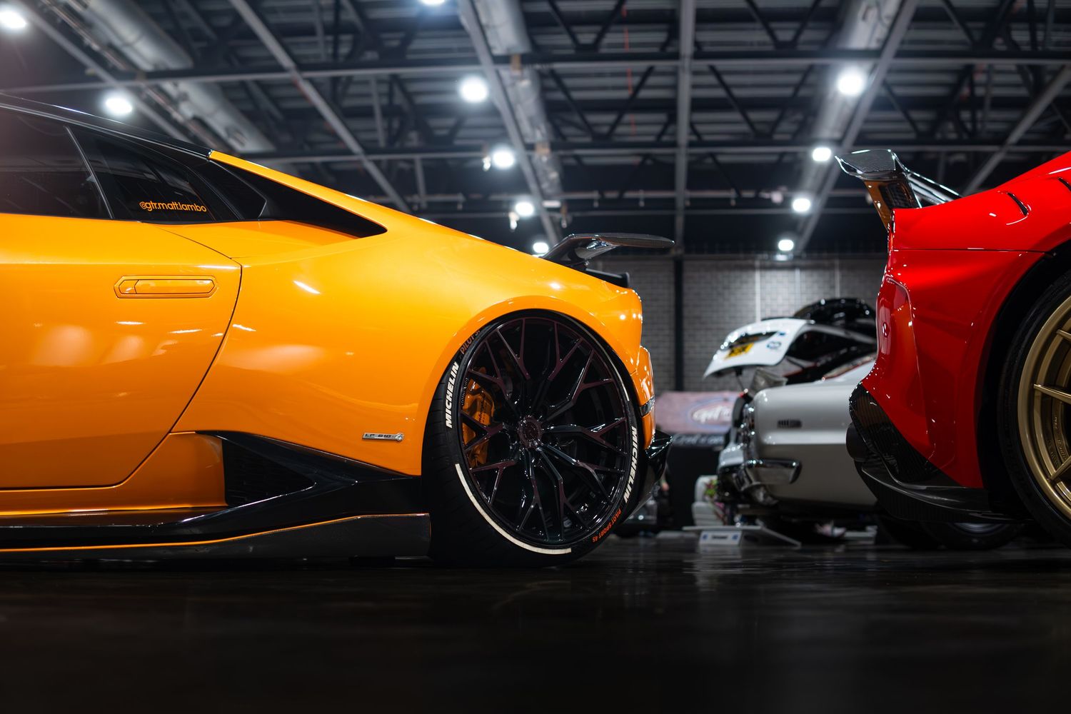 Orange Lamborghini Huracan beside a red Ferrari at an indoor automotive exhibition.