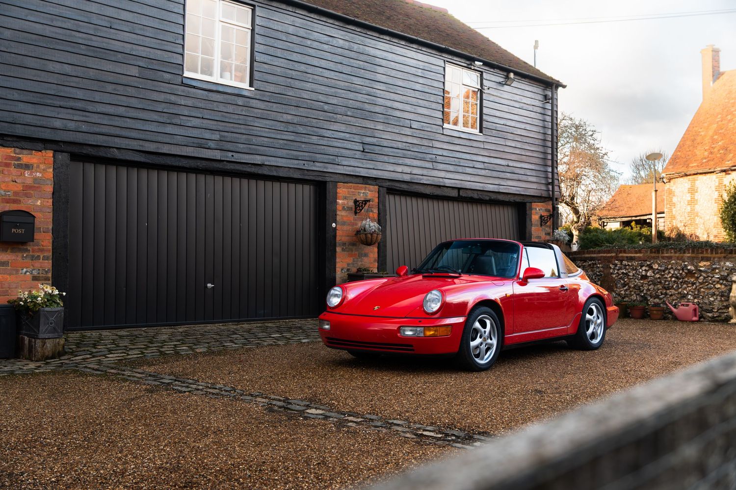 Classic red Porsche 911 Carrera 2 Targa parked outside a dark wooden garage in a rural English setting.