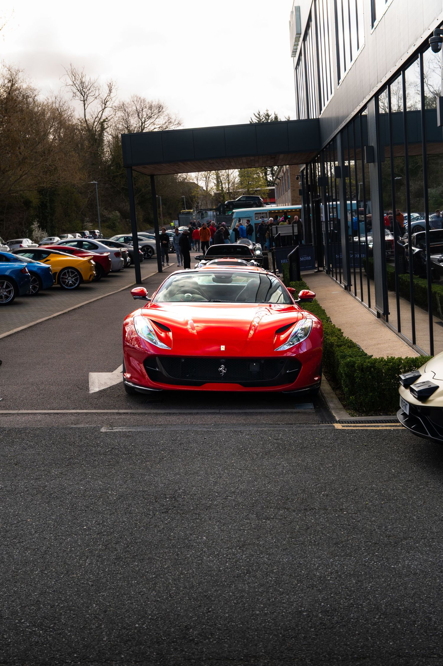 Red Ferrari 812 Superfast parked at entrance of modern automotive dealership with supercars behind.