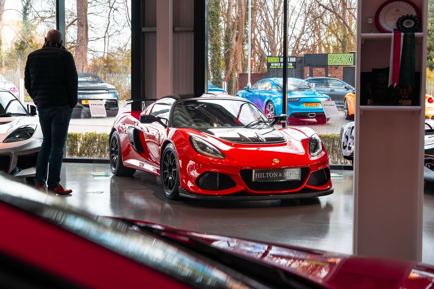 Red Lotus Exige Cup sports car on display inside a modern luxury car dealership showroom.
