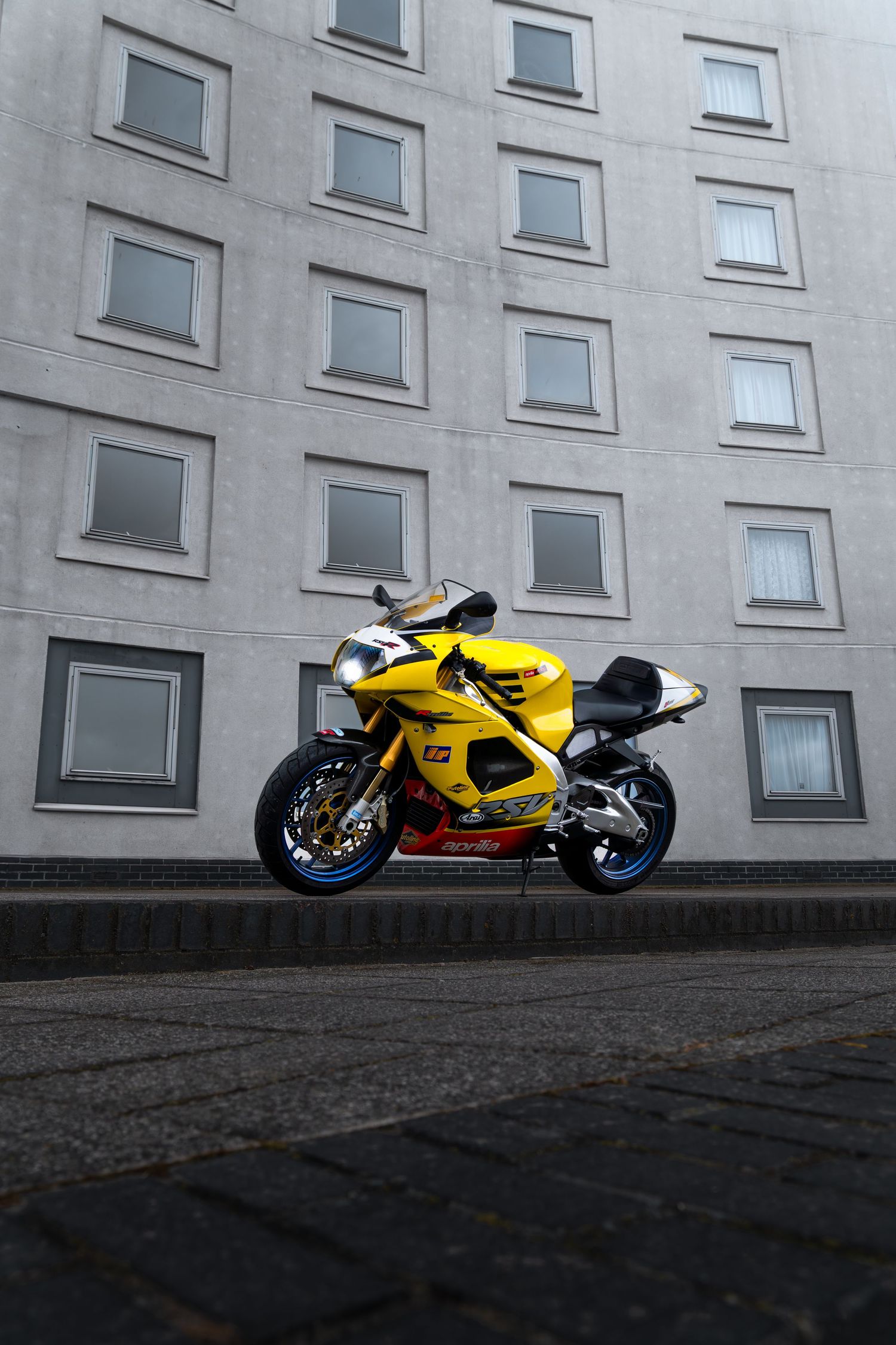 Yellow Aprilia superbike parked on cobblestones in front of a modern multi-story building.