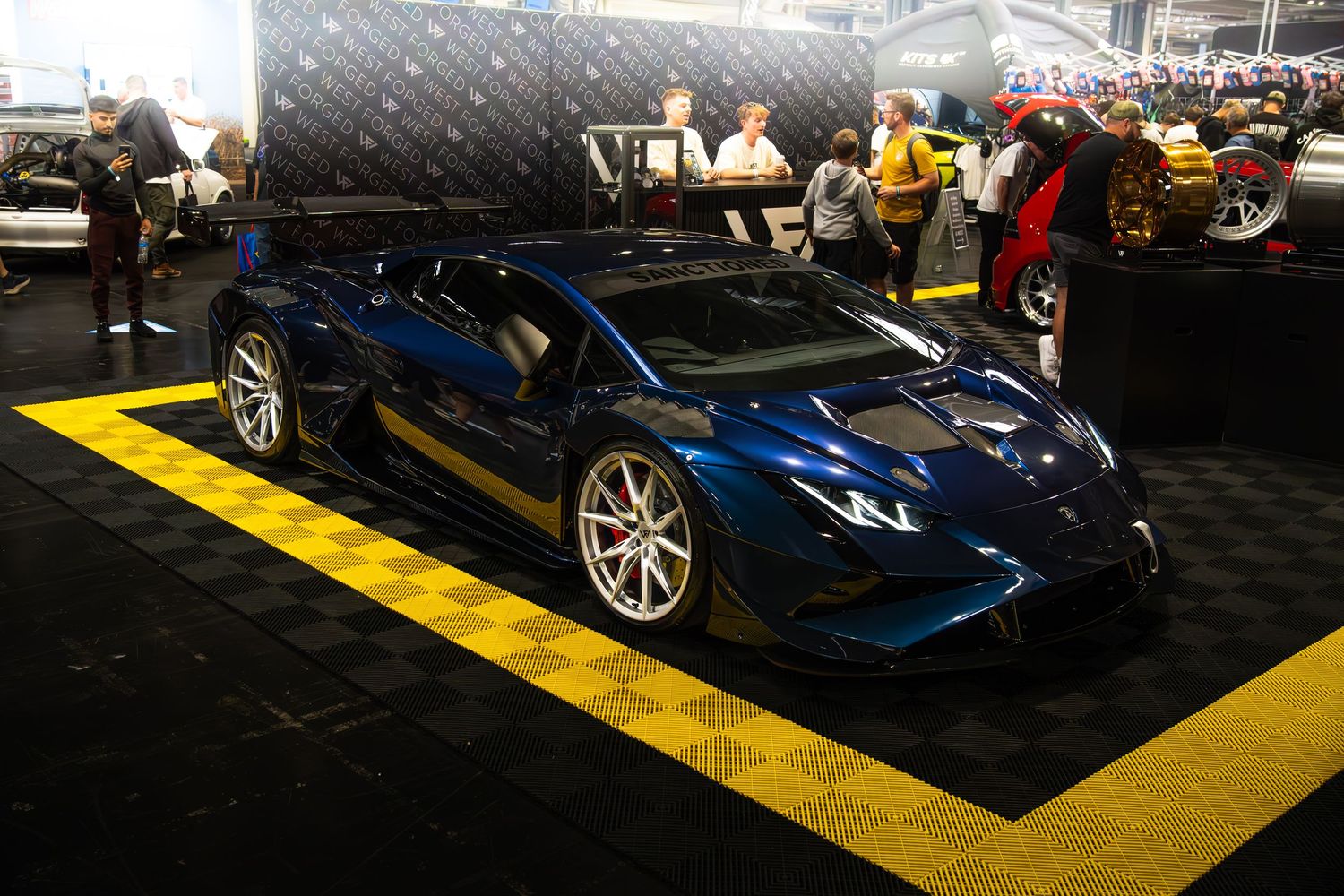 Dark blue Lamborghini Huracan with aerodynamic bodykit on yellow checkered flooring at a car show.