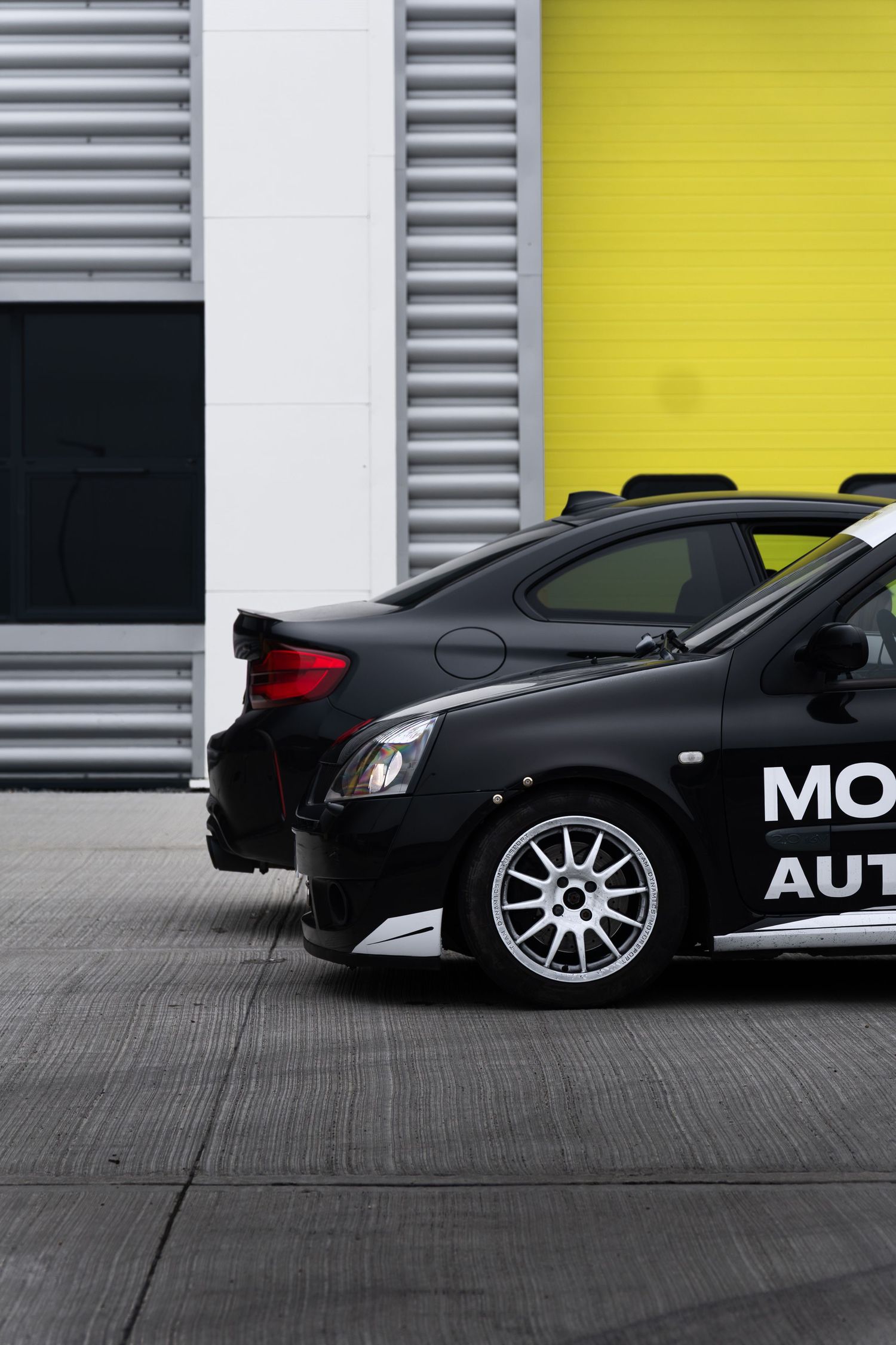 Close rear side view of a black Motion Automotive branded sports car parked against a yellow and grey garage door.