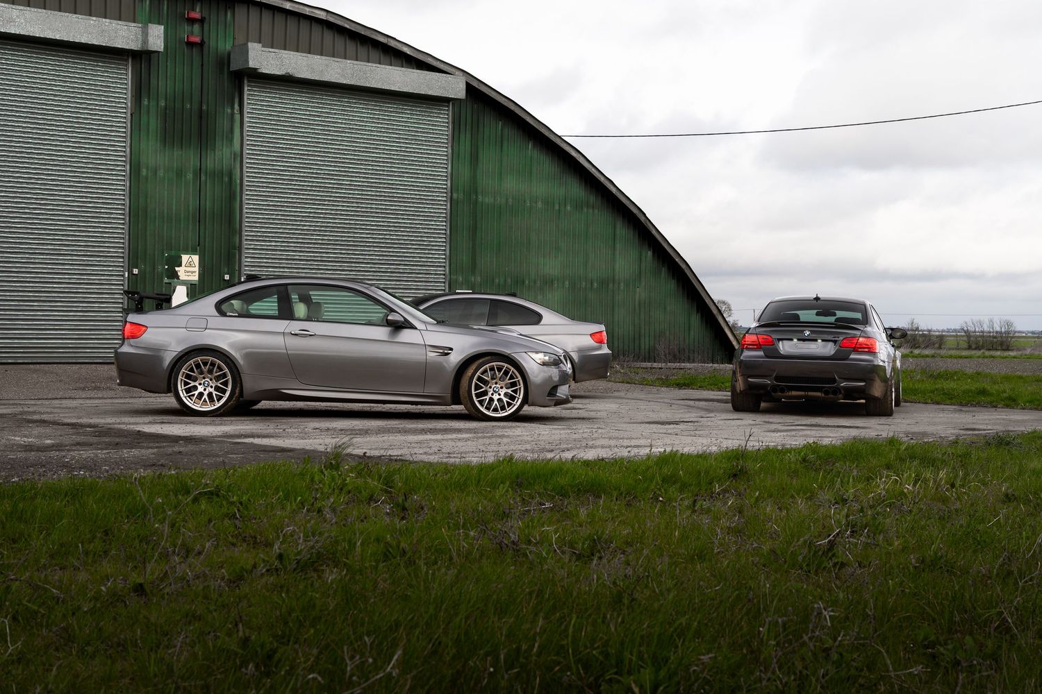 Gray BMW M3 E92 coupe parked beside a large curved green metal hangar building on grass.