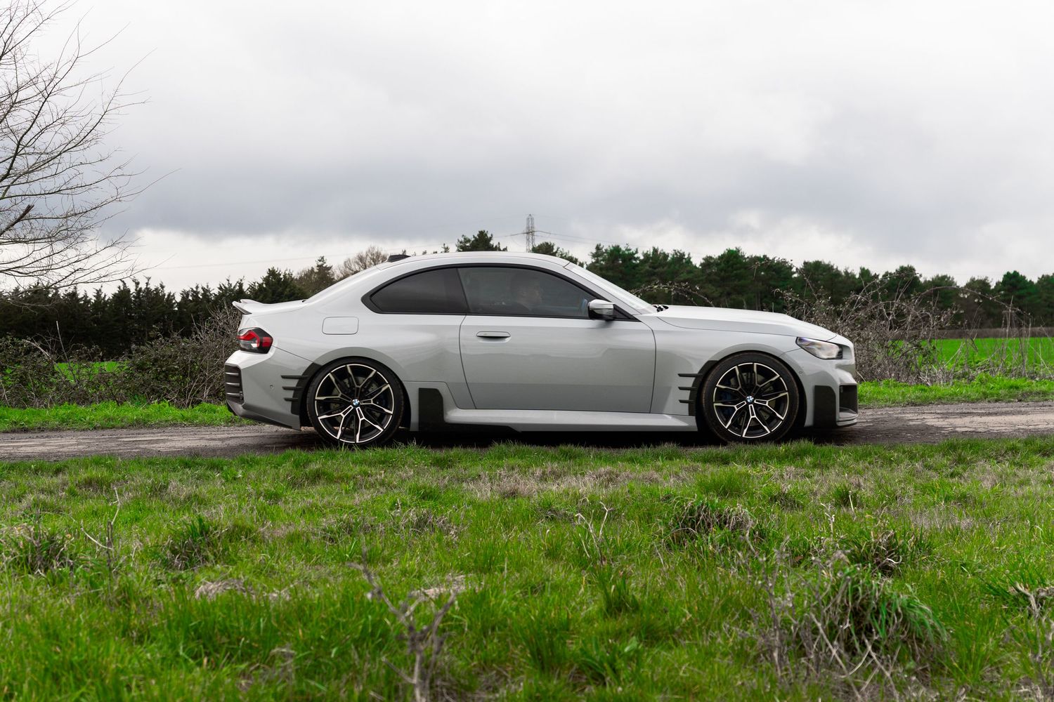 Silver BMW M3 E92 coupe parked on a country road surrounded by lush green grass under cloudy skies.