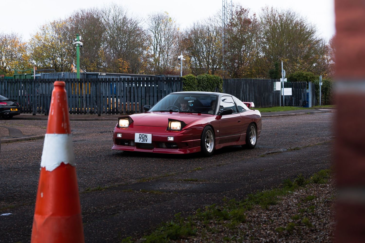 A red modified sports car drives past an orange traffic cone in an autumn car park.