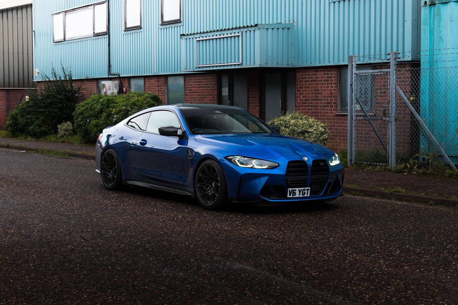 A blue BMW M4 coupe parked outside an industrial building on a cloudy day.