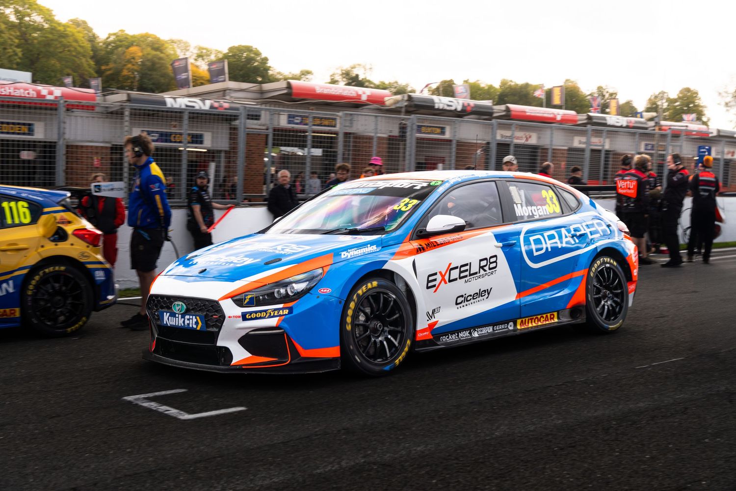 A Hyundai i30 N BTCC race car in blue Excelr8 livery sits in the pit lane at a motorsport event.