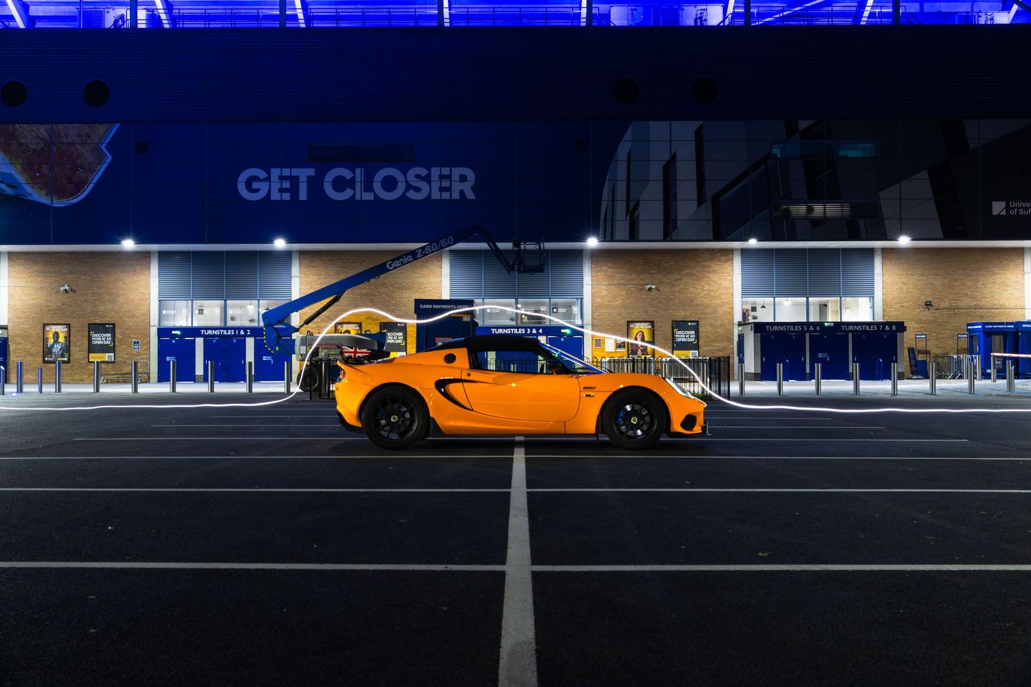 Side profile of orange Lotus Elise sports car parked at night outside blue-illuminated stadium GET CLOSER entrance.