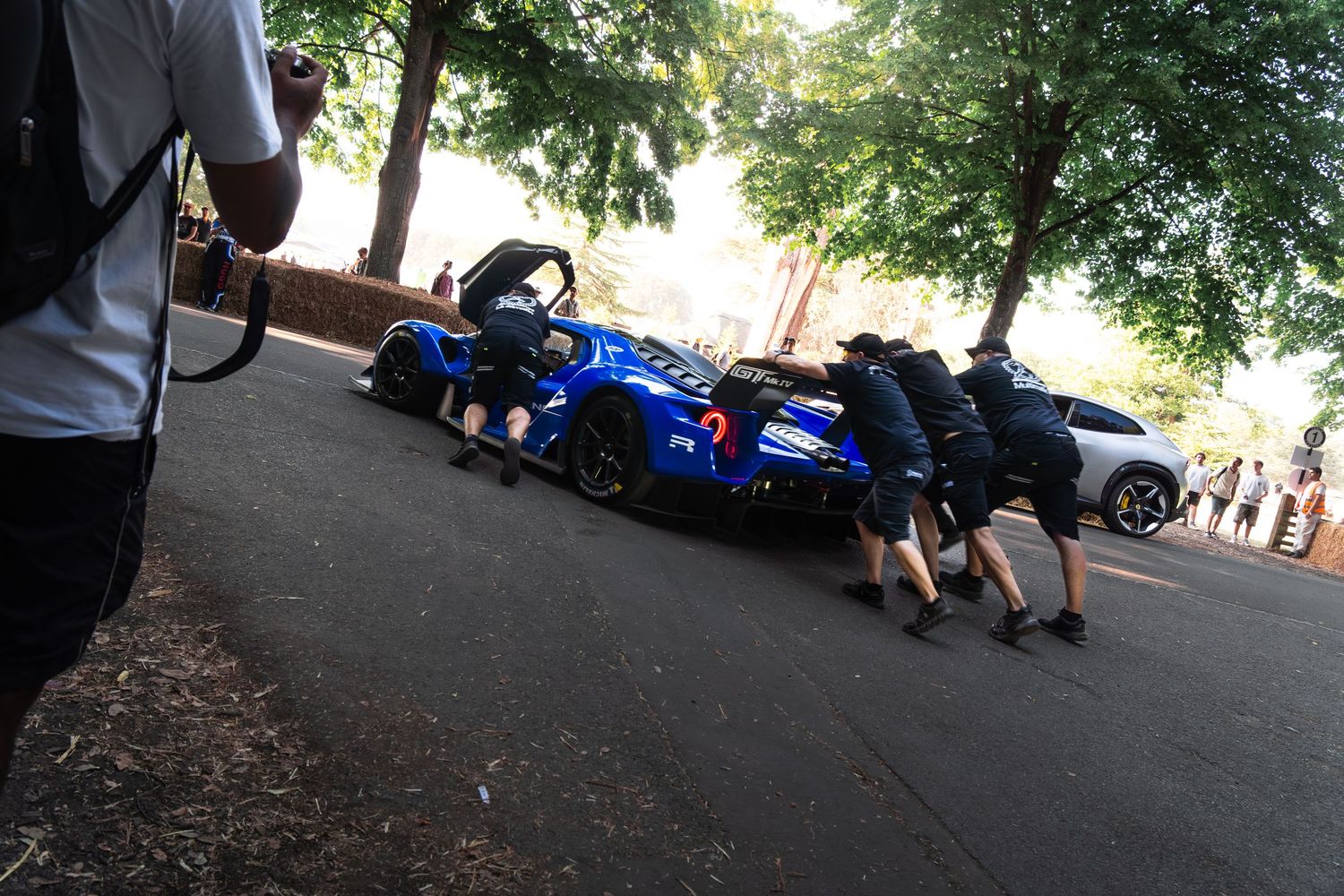Crew members pushing a blue racing sports car on a tree-lined track at an outdoor motorsport event.