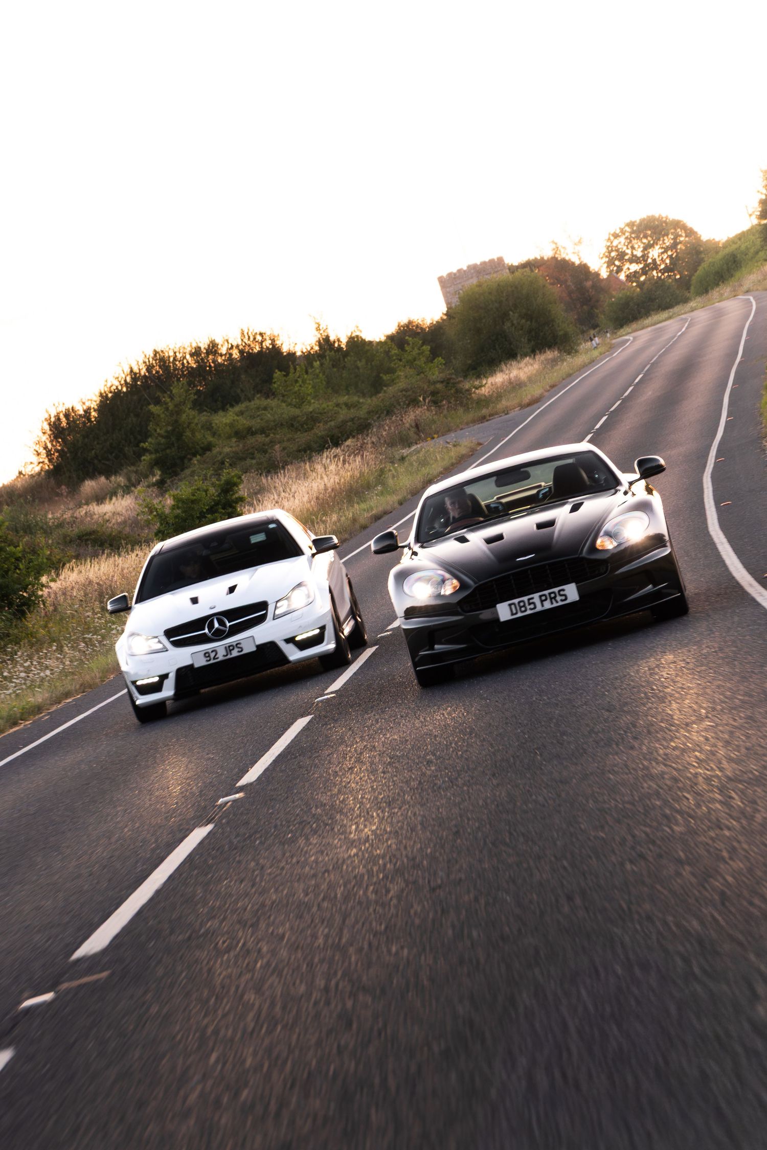 A white Mercedes-AMG and dark Aston Martin DBS race side by side on a country road at golden hour.