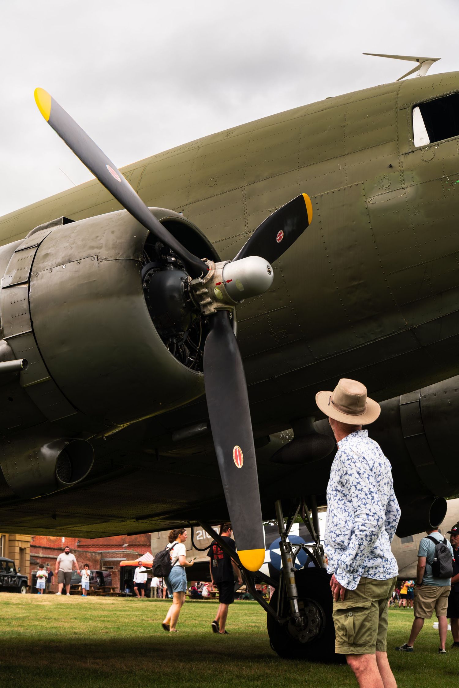 Man in straw hat admiring vintage WWII Douglas C-47 aircraft propeller at outdoor airshow on cloudy day.